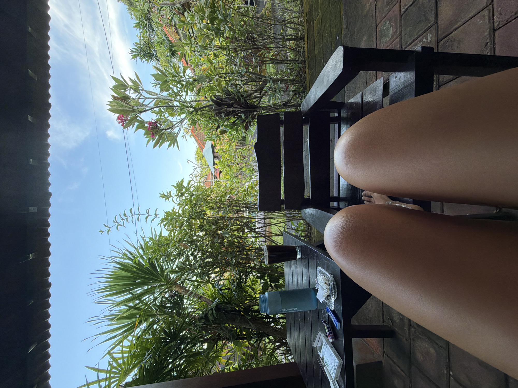 The image shows a partial view from a chair in what appears to be a garden or patio setting in Kutuh, Indonesia. Two legs of a person, tan in color, are visible. In the foreground, there is a dark wooden table with a blue water bottle, a small glass of dark liquid (possibly coffee or tea), an ashtray with cigarette butts, lighters, and a small package of items, possibly cigarettes. Behind the chair and table is lush green vegetation, including palm trees and flowering bushes. In the background, glimpses of buildings with red tile roofs are visible. The sky is blue with scattered clouds. A dark corrugated roof edge is visible in the upper left corner of the image. The entire scene is rotated 90 degrees counterclockwise.