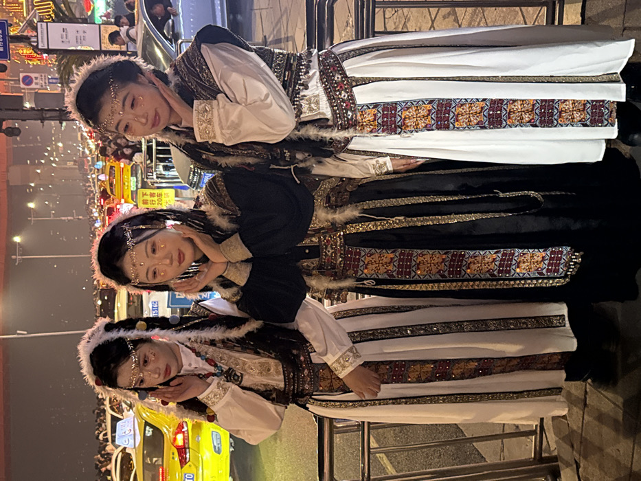 Three young women stand posed at night on a city street in Yuzhong District, China. All three are dressed in elaborate, traditional-style garments featuring long skirts with patterned and embroidered panels in gold, black, maroon, and white. They wear decorative vests over white or black long-sleeved blouses, adorned with metallic accents, beads, and feathers. Each woman has a delicate gold-colored headpiece across her forehead and small gold or glitter applications on her cheekbones and around her eyes. The woman on the left wears a white skirt and a vest with colorful beaded necklaces. The central woman wears a black skirt and top, with her hands cupped near her chin. The woman on the right wears a white skirt and a vest, also posing with her hands cupped. Their expressions are serene. Behind a metal barrier in the immediate background, a busy street is visible with heavy traffic, predominantly yellow taxis, and other vehicles with their headlights illuminated. Distant city lights and streetlights illuminate the urban environment under a dark sky.
