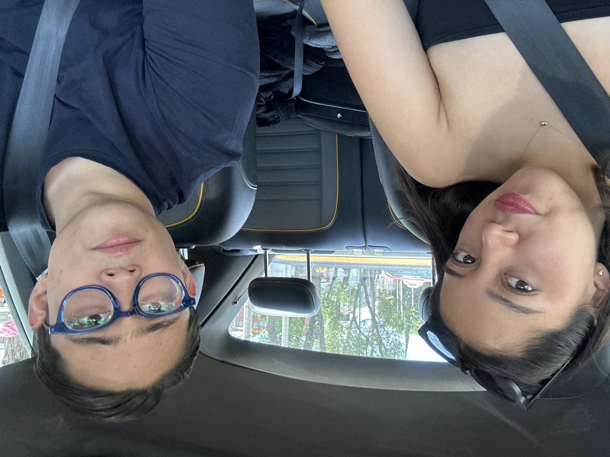 Two individuals, a male and a female, are seated in a car wearing seatbelts. The man, on the left, wears glasses and a navy blue shirt. The woman, on the right, wears a black top and has sunglasses atop her head. They are positioned in front of black leather seats with yellow accents. Through the car window, trees and buildings can be seen. The reflection in the car window shows details of Mexico City, Mexico. Taxis are visible in the window and parked cars on the street.