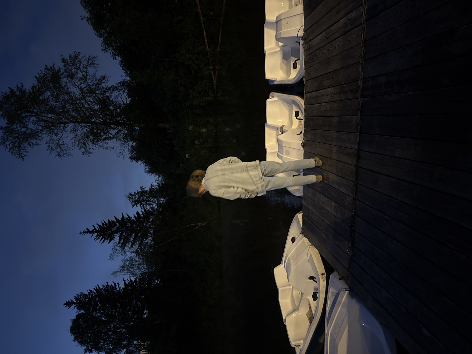 A person stands on a wooden dock at night, facing away from the camera. They are wearing a light-colored hooded sweatshirt and blue jeans. In front of them, several white pedal boats are docked. The dock is made of dark wood planks. The background consists of a dark body of water, with a tree line and the night sky visible beyond. The scene is dimly lit, suggesting it is either dusk or dawn. This location is the beach area of the Izhgorskoye Reservoir in Izhevsk, Russia.