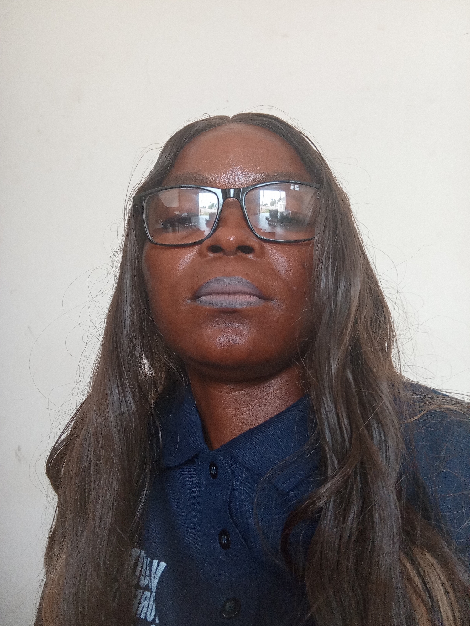 A medium shot shows a dark-skinned woman with long, dark brown hair and glasses. She is wearing a dark blue collared shirt with buttons and a logo on the left side of the chest. The background is a plain, light-colored wall. The woman is looking directly at the camera.