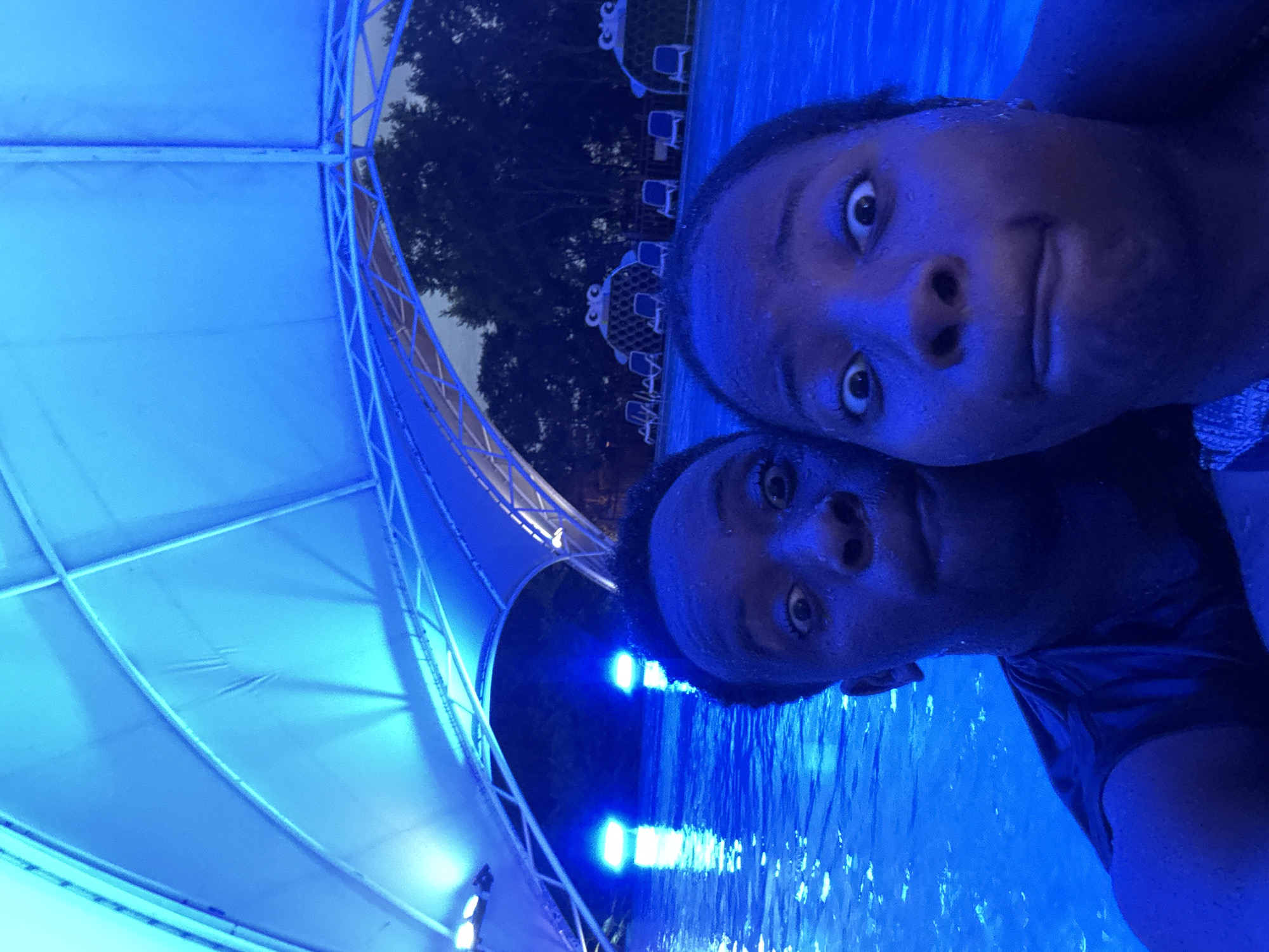 Two individuals are submerged in a swimming pool under a large blue tent structure at night. The water is illuminated with bright blue lights, reflecting off the surface. In the background, trees and outdoor seating areas with lounge chairs and cabanas are visible. The individuals are looking at the camera with exaggerated expressions.