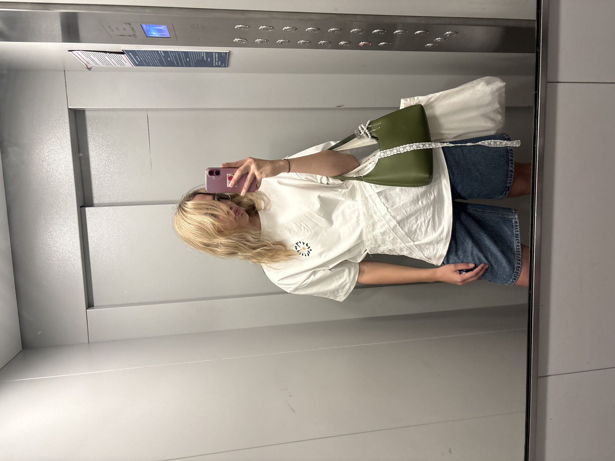 An individual, appearing to be female with blonde hair, stands in an elevator and takes a mirror selfie. She is wearing a white t-shirt with a small graphic, denim shorts, and is holding a green handbag. A white tote bag is also visible. The elevator has gray walls and a metallic panel with buttons on the right side. The scene is consistent with an elevator in a residential or commercial building. There is no information in the image to suggest the location is Krasnoyarsk, Russia.