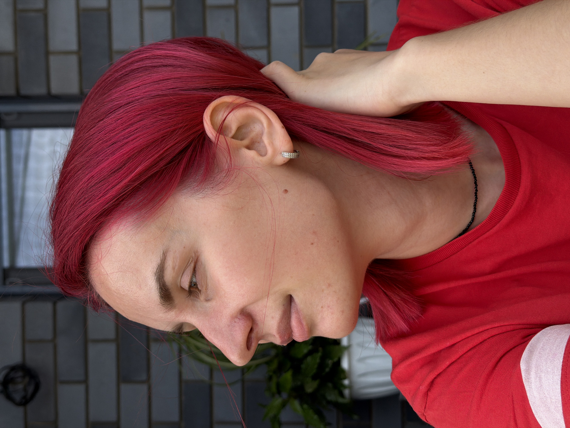 A medium shot captures a young Caucasian woman with bright red hair, her head turned to the left in profile. She is wearing a red t-shirt with a white stripe on the sleeve and a thin black beaded necklace. Her right hand is raised to the back of her head, with her fingers gently touching her hair. Her left hand is not visible. She is wearing a small, sparkly hoop earring in her left ear.
The background consists of a brick wall, appearing to be dark grey or black. To the left of the woman, a large potted plant with green leaves is partially visible, suggesting an outdoor setting. The lighting appears to be natural daylight.
The provided context mentions the location as Chaikovsky, Russia. Without further information or visual cues within the image, it is impossible to definitively confirm this location or provide specific details about the city itself. The image depicts a private moment, and there are no public landmarks or distinctive features that would place it within a specific urban environment.