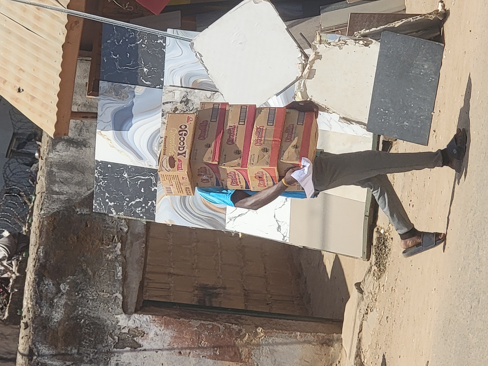 A man carries tiles and multiple boxes of Goody Goody Krunch drink on his head. He wears grey pants, a blue shirt and sandals. The location appears to be an exterior area in Maiburiji, Nigeria, with a partially visible building and corrugated metal structure in the background. The area has weathered walls, a concrete road, and discarded debris.