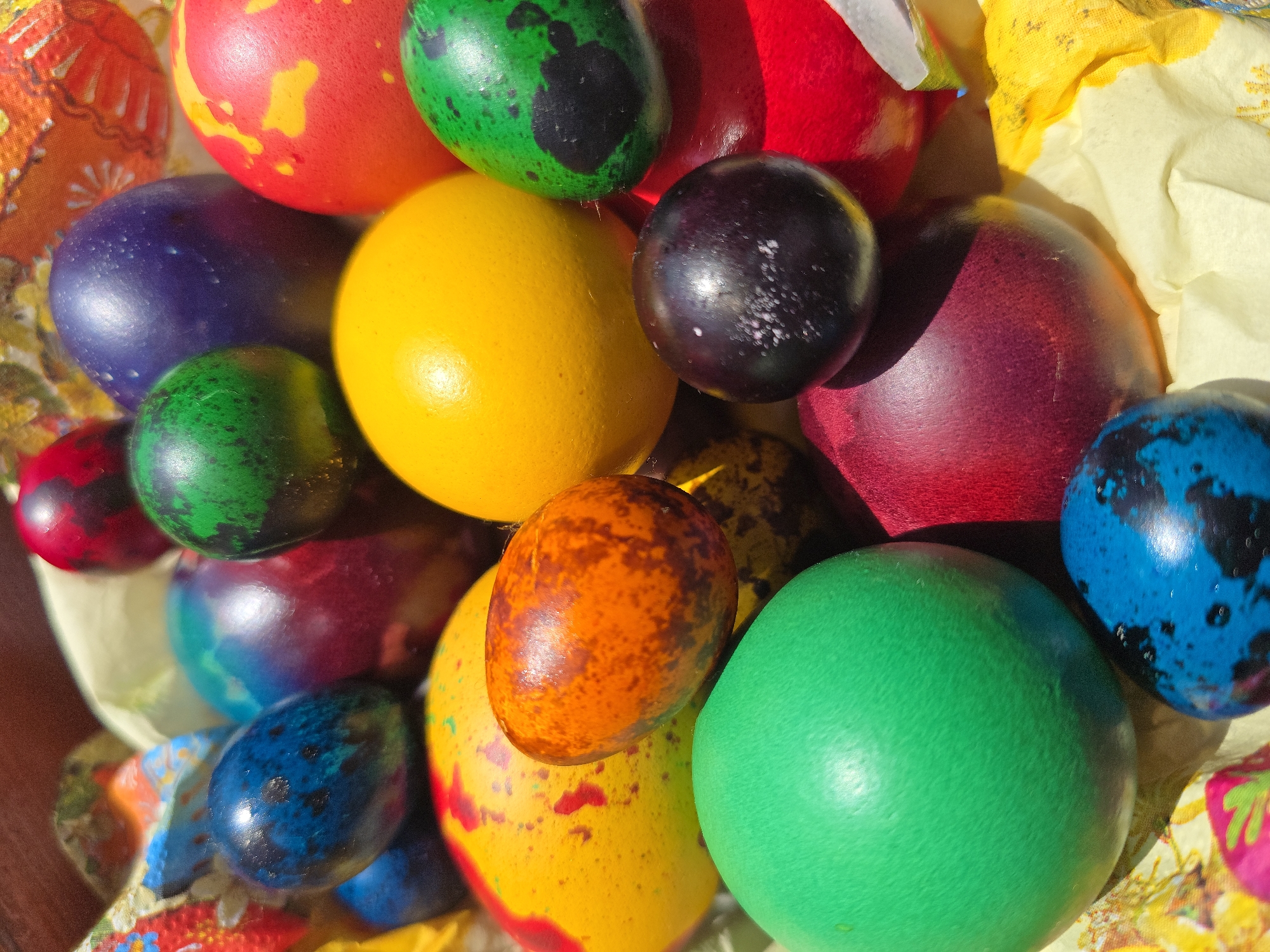 A close-up shot reveals a basket filled with vibrantly colored Easter eggs. The eggs come in various shades, including red, yellow, orange, green, blue, purple, and black, some featuring speckled or mottled patterns. The eggs are nestled among crumpled white and yellow paper, and part of a colorful, patterned fabric can be seen around the edges of the basket. The lighting is bright, suggesting an outdoor setting with direct sunlight illuminating the scene. This likely takes place during Easter festivities, although no location in Varna, Bulgaria is determined from this image.