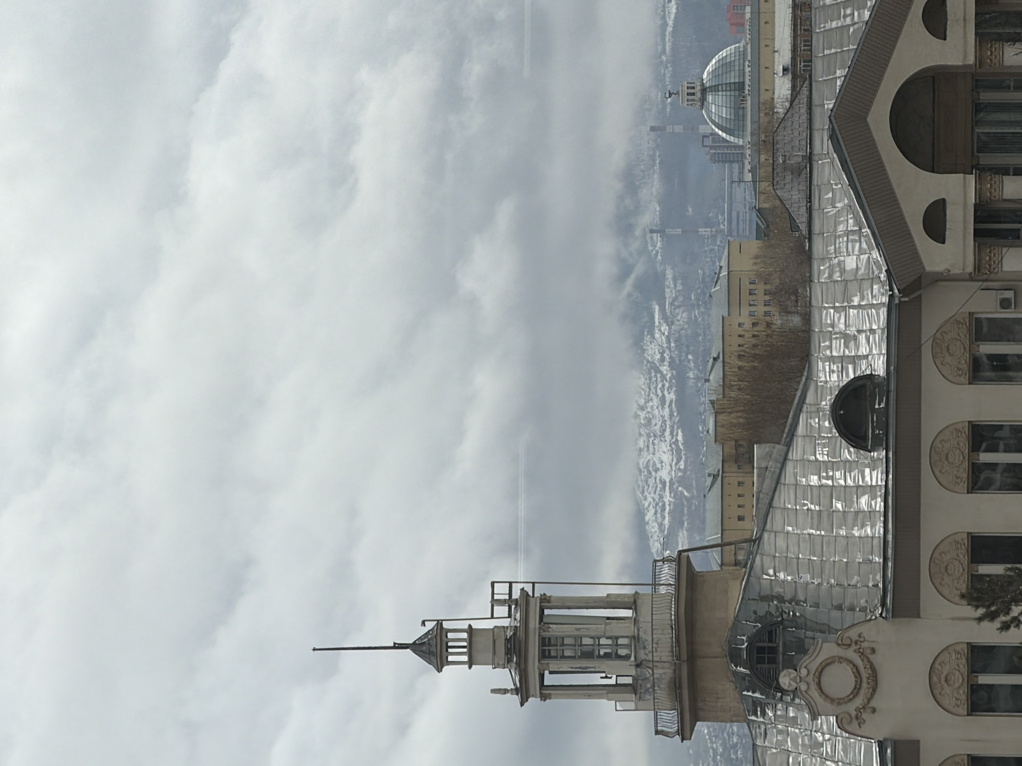 The image shows a building in Krasnoyarsk, Russia, with distinctive architectural features including a turret and a curved roof covered with reflective metal tiles. In the background, a city landscape with buildings, including one with a dome, is visible. The sky is overcast with clouds, and there appears to be snow on distant hills. The architecture is ornate, with decorative circular elements on the façade of the building.