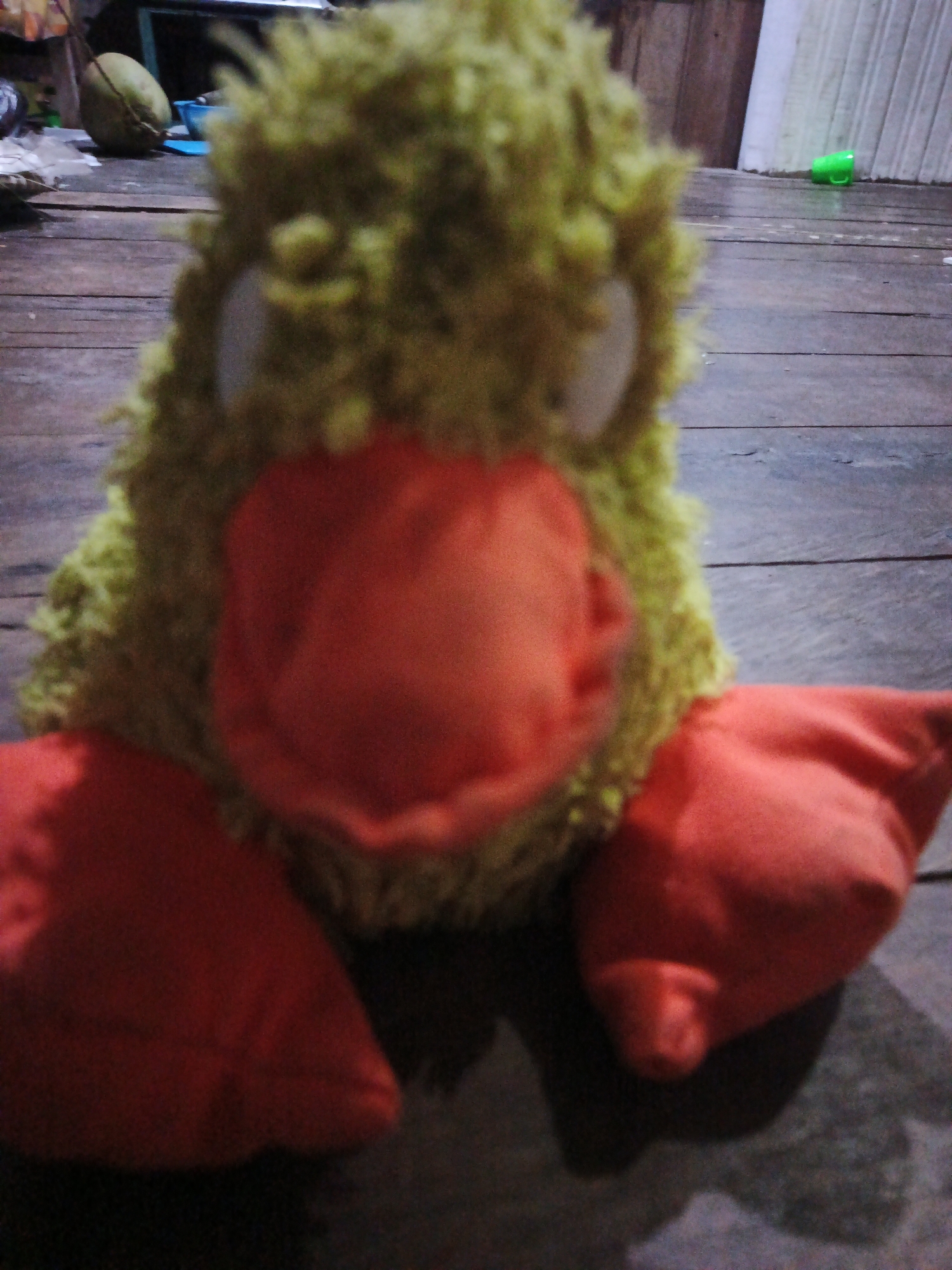 A close-up, slightly out-of-focus image shows a fluffy, lime-green stuffed duck with large orange feet and a prominent orange beak. The duck is positioned on a weathered wooden floor. In the background, out of focus, are various objects on a wooden surface, including a coconut, a blue bowl, and a small green object. The scene appears to be indoors or in a covered outdoor area with a wooden wall in the background. The lighting is dim, casting shadows. The image is undated and its specific location in Sinar Bulan, Indonesia, is not visually discernible from the provided image.