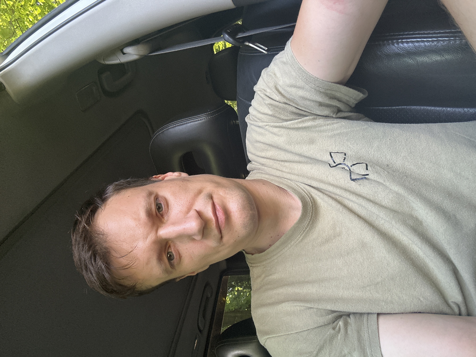 A man is sitting in the driver's seat of a car, looking at the camera. He is wearing a light brown t-shirt with a black graphic on the left chest. The car interior is visible, including the black leather seats, the sunroof, and the interior trim. Green trees and foliage are visible through the car windows, suggesting an outdoor setting, possibly in a wooded area. The specific location of Shatava, Ukraine, cannot be determined from the visual information alone.