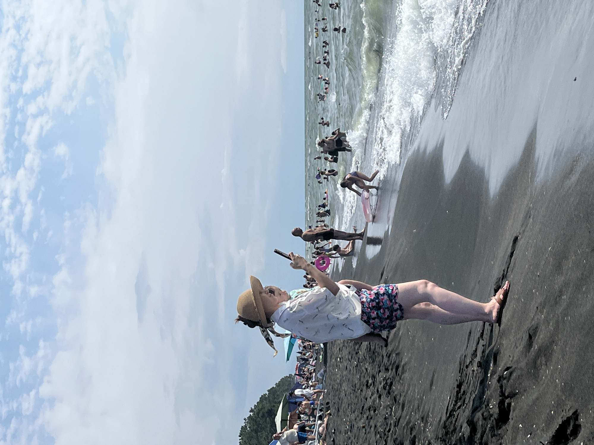 The image depicts a crowded beach scene on a cloudy day, likely in Ureki, Georgia, a resort town known for its magnetic black sand beaches. The foreground is dominated by the dark, coarse sand, with a woman standing on it. She is wearing a wide-brimmed hat, a patterned shirt, and floral shorts, and is holding a child and a smartphone. Her focus is on the phone.
The midground shows the edge of the water with gentle waves breaking on the shore. Several people are visible in the water, including children playing and adults swimming. A pink inflatable ring is visible near the water's edge, and a young girl in a blue swimsuit is interacting with it.
The background is filled with a multitude of people scattered across the beach and in the sea, suggesting a popular tourist spot. Beach umbrellas and trees line the distant shore. The sky is a mix of blue and white clouds. The overall impression is one of a lively and populated summer day at the beach.