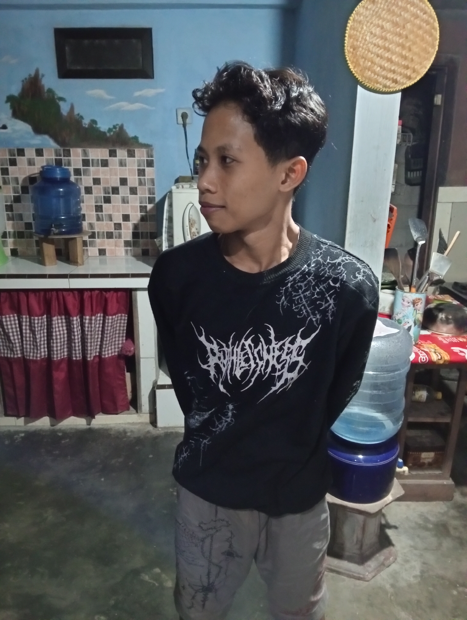 The image shows a young male standing in what appears to be a kitchen or utility area, possibly in Trenggalek, Indonesia. He is wearing a black long-sleeved shirt with white, graphic designs and light-colored pants. The room has light blue walls, a tiled backsplash, and red and white checkered curtains hanging below a counter. In the background, there is a large blue water dispenser, a woven circular object hanging on the wall, and shelves with various items. The lighting suggests an indoor setting, possibly at night.