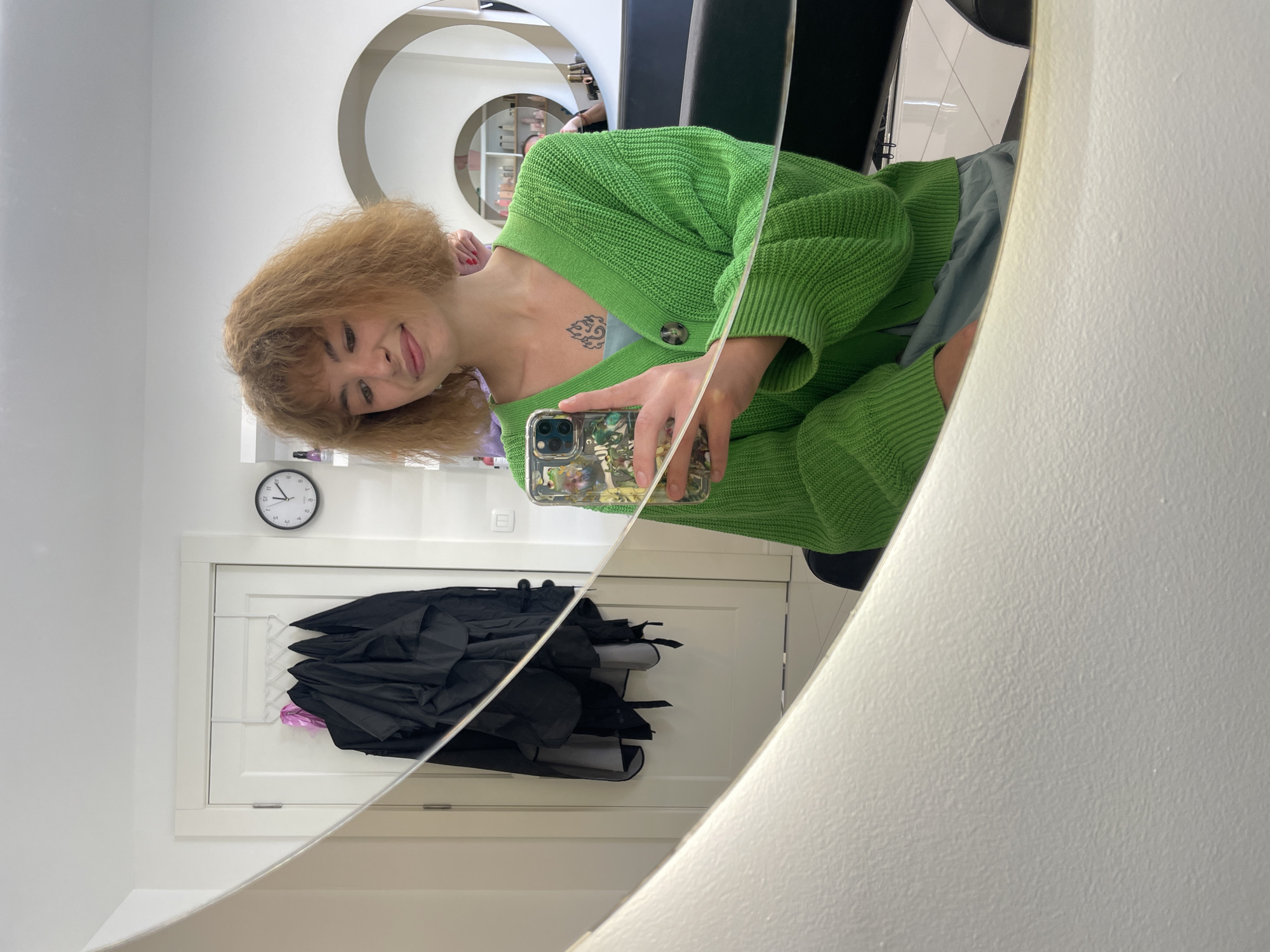 A woman is taking a selfie in a salon, likely in Rafailovići, Montenegro, based on the context clues. She has shoulder-length blonde, curly hair and is wearing a green cardigan over a light blue top. A tattoo is visible on her chest. She is holding a smartphone with a patterned case. The reflection in the mirror shows a salon environment with a white wall, a round mirror, and salon furniture, including a black chair. Behind the woman in the reflection is a closed white door with black umbrellas hanging inside. A clock is mounted above the door. The image is taken at an angle due to the selfie perspective.