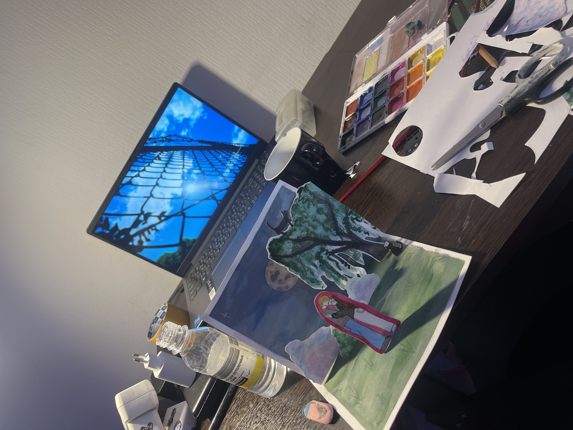 The image depicts a cluttered desk with various artistic and technological items. A laptop with a screensaver featuring a blue sky and a wire fence is visible on the left. A partially visible black mug sits next to it. A watercolor palette, a container, a pair of scissors, and paper cutouts are to the right of the mug. At the bottom right, a bottle of clear liquid is placed near an Apple charger, tape, and other smaller items. The focal point is a painting or drawing of a couple under a tree, seemingly set in a night scene with a moon. An eraser rests beside the artwork.
There is no information about Saint Petersburg, Russia, in the given image.