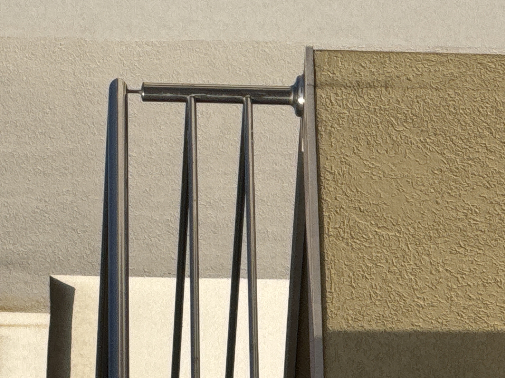 This is a close-up, outdoor, eye-level shot focusing on the texture of a stucco wall and a metal railing. The wall, located on the right side of the frame, is a light tan color with a rough, stucco-like texture. A shadow is cast on the lower portion of this wall, suggesting it is daytime. To the left of the stucco wall, a portion of a building with a lighter, smoother texture is visible, acting as a backdrop.
In the foreground, a metal railing system is prominent. It consists of several thin, vertical metal bars and a thicker horizontal bar connecting them at the top. The metal appears to be polished or brushed stainless steel, reflecting the ambient light. The railing is attached to the stucco wall on the right with a circular fitting. On the left, another vertical metal element, part of the railing structure, is visible. Between the vertical bars of the railing, a lighter surface is visible, possibly another part of the building's architecture.
There are no people or animals visible in the frame. The overall scene suggests an exterior architectural detail, possibly a balcony or staircase railing, in a location with a stucco-finished building. The lighting indicates it is likely daytime, with sunlight creating the shadow. No text is visible in the image. The provided location context of Budva, Montenegro, suggests this could be a common architectural element found in that region.