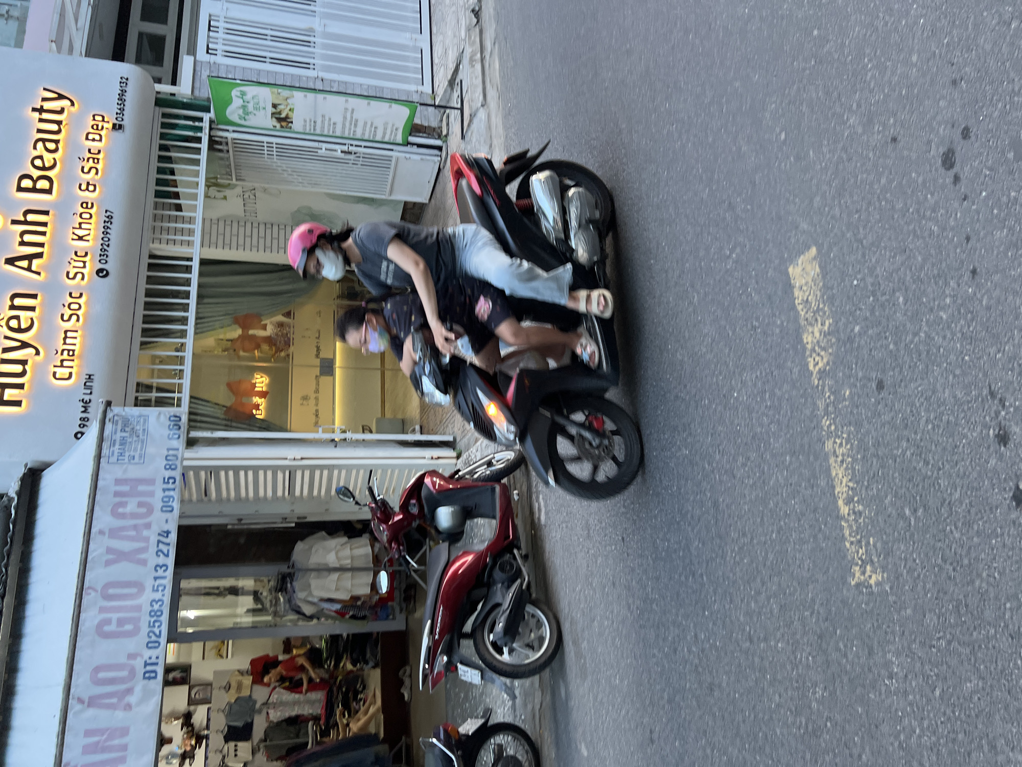 In Nha Trang, Vietnam, an image captures a street scene with several motorcycles and a storefront. The storefront displays a sign for "Huyen Anh Beauty," offering "Chăm Sóc Sức Khỏe & Sắc Đẹp" (Health Care & Beauty Services) with phone numbers 0392099367 and 0365896132. Another sign above a shop reads "QUẦN ÁO, GIỎ XÁCH" (Clothes, Bags) with phone numbers ĐT: 02583.513 274 - 0915 801 660. Inside the "QUẦN ÁO, GIỎ XÁCH" shop, clothing items are visible on display racks.
Two individuals are riding on a black scooter. The person in the front, a woman, wears a pink helmet, a grey t-shirt, and light-colored pants, and a white mask. The person behind her, a child, wears a dark patterned t-shirt and a light-colored mask. They are both wearing sandals. A red scooter is parked next to the storefront. Another black scooter is partially visible on the left side of the image. The street is paved with asphalt and has a yellow line marking. The overall setting appears to be a commercial street with shops and pedestrian activity.