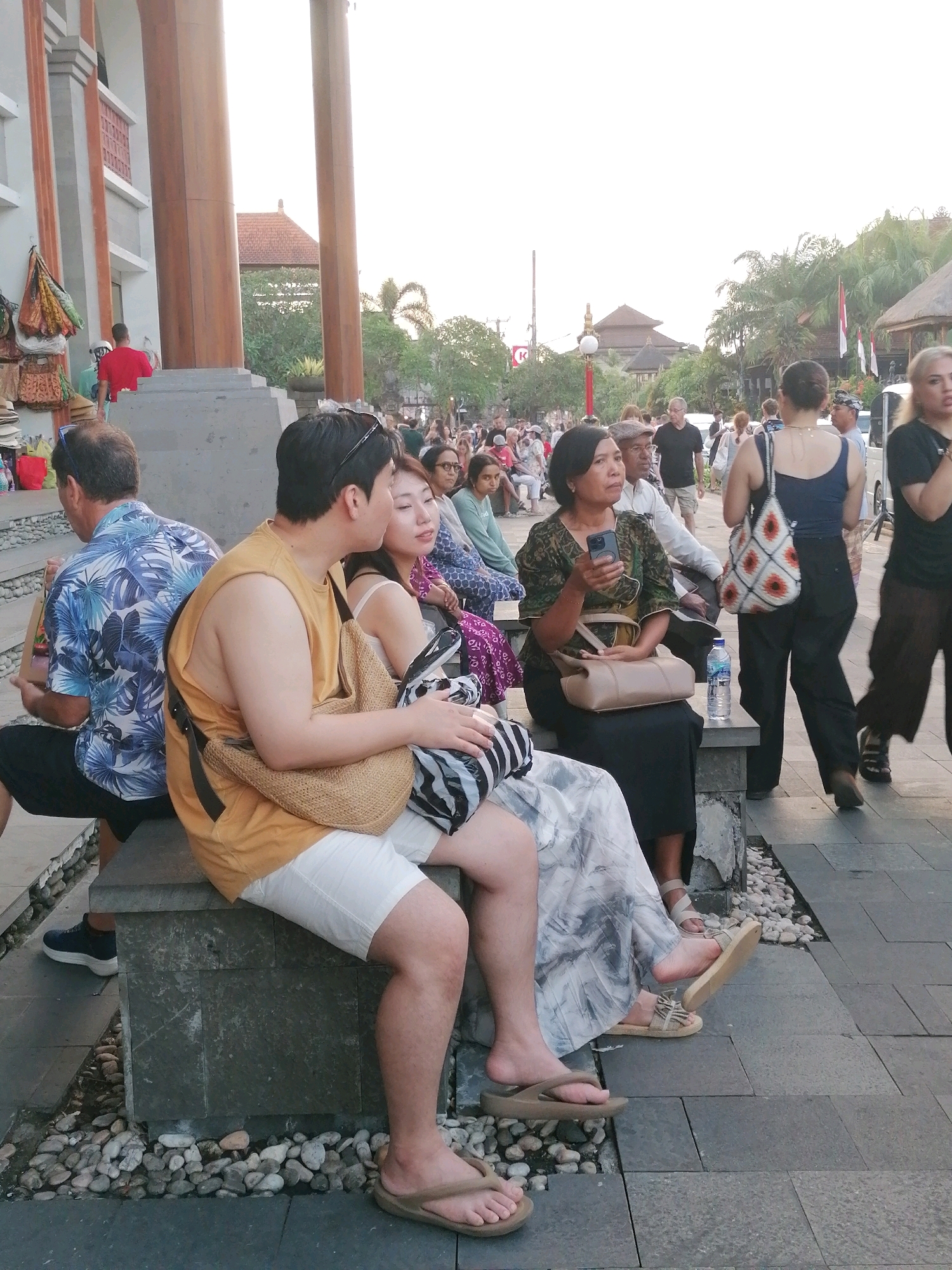 In the city of Ubud, Indonesia, a group of people are seen sitting and standing on a paved walkway. In the foreground, a man wearing an orange sleeveless shirt and white shorts sits on a stone bench, facing away from the camera. Beside him, a woman in a patterned dress is also seated. To their left, another man in a blue and white floral shirt and black shorts is seated.
Behind the seated individuals, a crowd of people are visible, some walking and others standing. One woman in a green patterned top holds a smartphone. A woman in a blue tank top and black pants walks away from the camera, carrying a patterned bag. Another woman, partially visible, wears a black top and brown pants.
The environment appears to be an outdoor public space, possibly a marketplace or a tourist area, with buildings and palm trees in the background. The lighting suggests it is late afternoon or early evening.