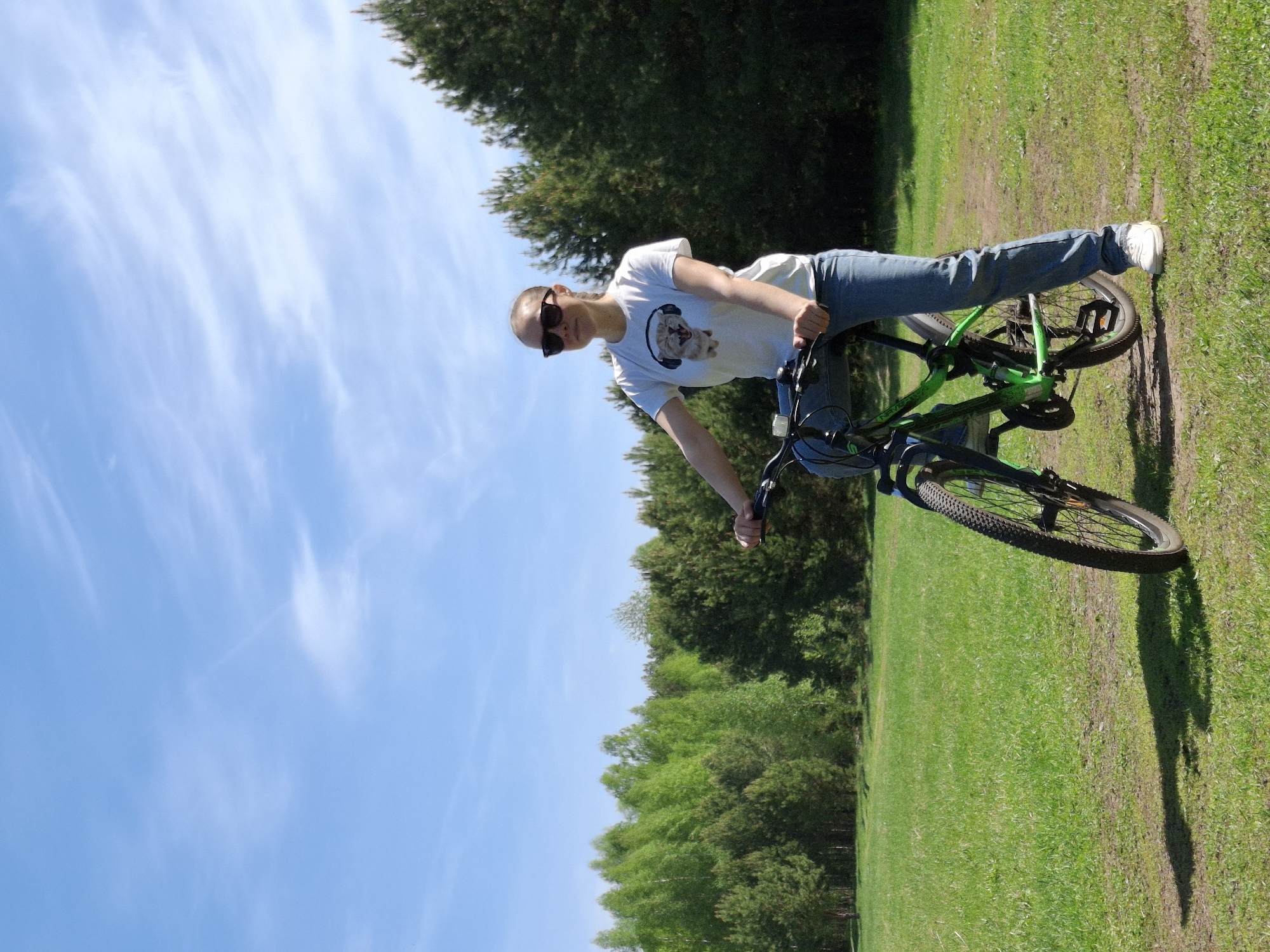 A fair-skinned person wearing sunglasses, a white t-shirt with a graphic, and blue jeans stands next to a green and black mountain bike. The bicycle rests on green grass. Behind the person and bicycle is a row of trees and a blue sky with wispy clouds. The scene appears to be outdoors in a park-like setting. The location is identified as the city of Chaikovsky, Russia.