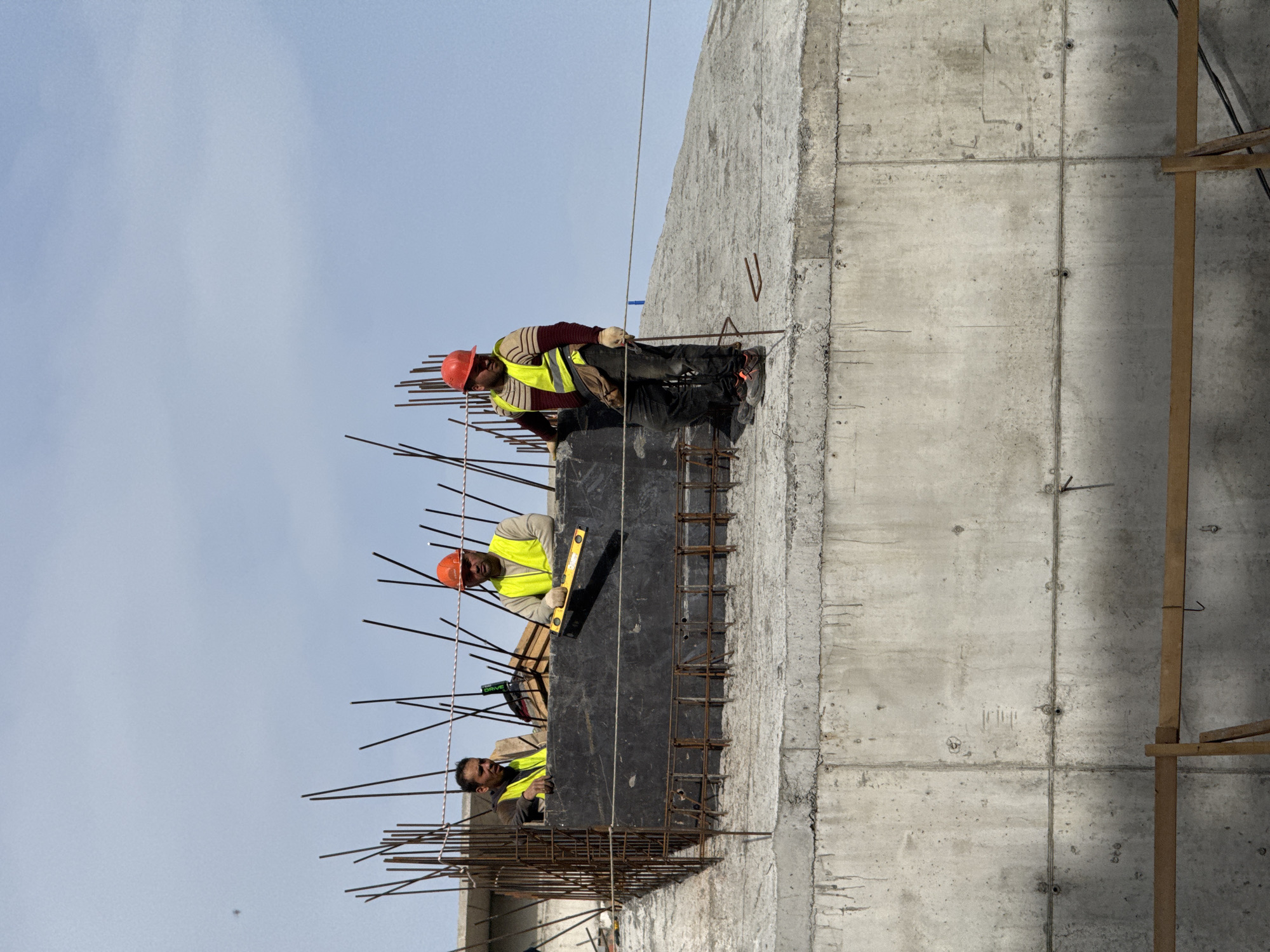 Three construction workers are working on the outside of a concrete building in Kazan, Russia. The workers are wearing safety vests and hard hats. One worker is using a level to check the alignment of a black form. The workers are standing on a rebar platform attached to the side of the building. The sky is overcast.