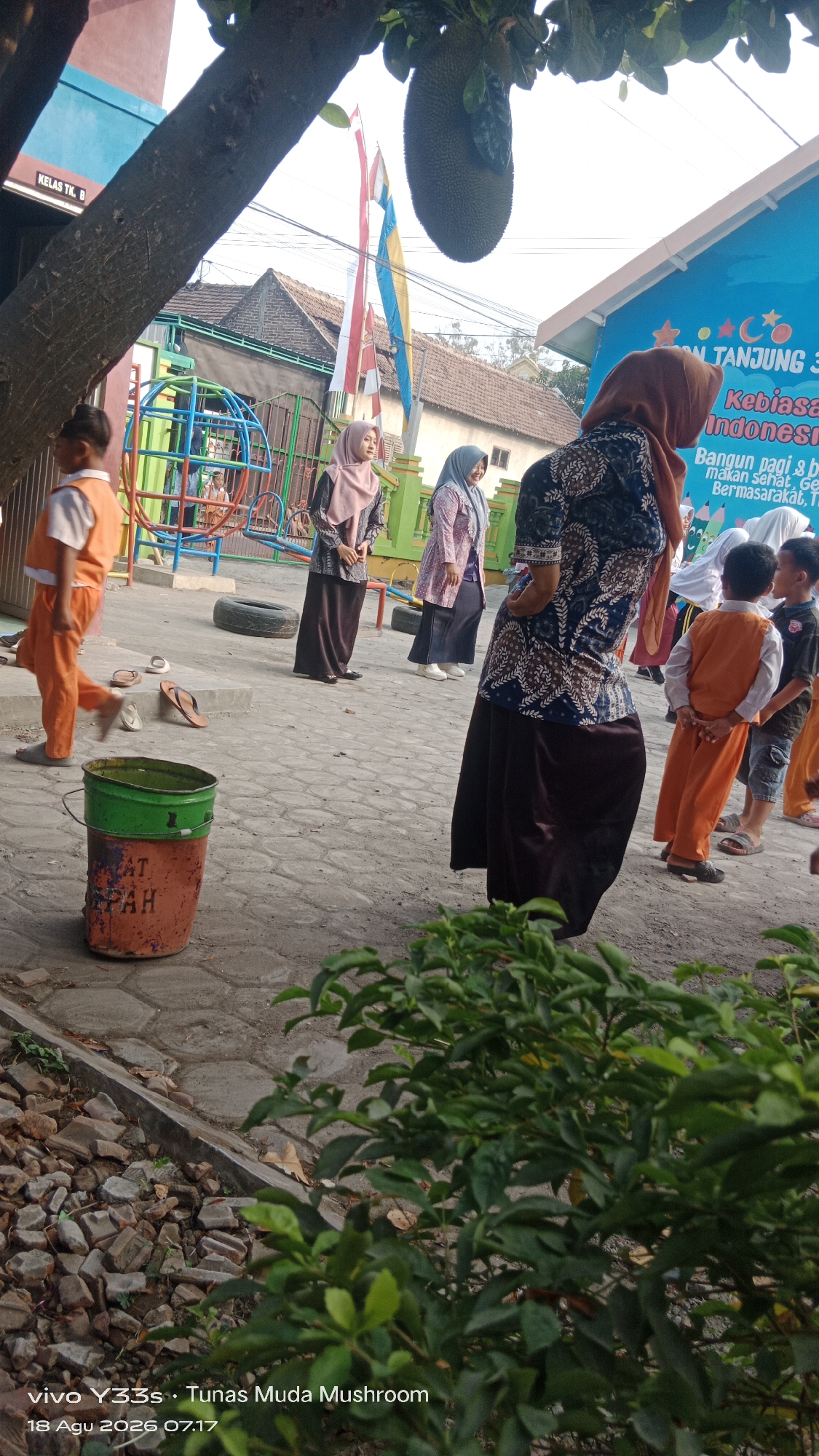 The image depicts an outdoor scene at what appears to be a school or community center in Tanjung, Indonesia. In the foreground, a large tree branch with a jackfruit hangs overhead. The ground is paved with interlocking concrete blocks and there are scattered bricks and rocks to the left. A green and orange painted bucket with the word "SAMPAH" (Indonesian for "trash") is visible on the ground.
Several people are present, mostly women and children. A young boy in an orange uniform is walking from left to right. Two women in headscarves and traditional Indonesian attire, possibly teachers, are standing and observing the scene. Another woman in a patterned blue and white outfit with a headscarf is also present, facing away from the camera. Several children in orange uniforms are interacting with each other. Behind them, a colorful playground structure with blue and green elements is visible. In the background, there are buildings with red and blue accents, and Indonesian flags flying. A blue building in the background has text in Indonesian that includes "TANJUNG", "Kebiasaan", "Indonesia", and "Bangun pagi". The overall atmosphere suggests a sunny day with a group of people engaged in an outdoor activity.