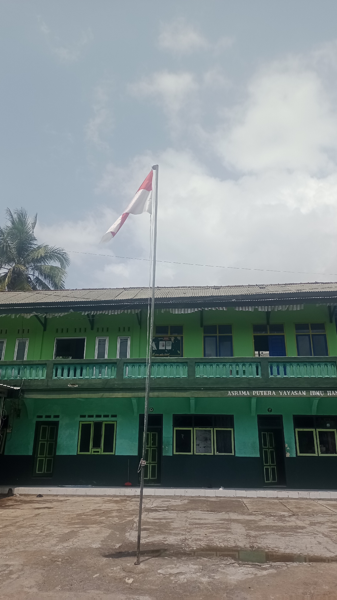 The image shows a two-story building with a green facade and a dark grey corrugated metal roof. A flagpole stands in the foreground, with a red and white Indonesian flag flying from it. The building appears to be a school or dormitory, with the text "ASRAMA PUTERA YAYASAN IBNU HAN" visible above the lower level windows. The lower level has several doors and windows, all with dark green frames. The upper level features a balcony with a green railing and multiple windows. A palm tree is visible on the left side of the building. The ground in front of the building is a concrete courtyard, with some areas appearing wet, possibly from rain. The sky is partly cloudy. The scene is located in Karyamandala, Indonesia.
