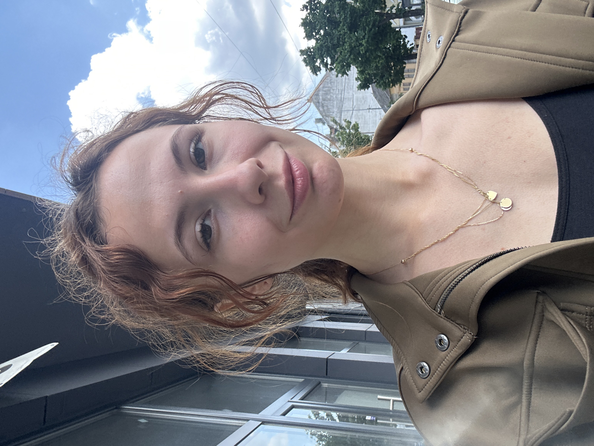 A selfie taken outdoors with a woman as the main subject. She has wavy, reddish-brown hair, light skin, and is wearing a black top and a brown jacket. Around her neck is a gold necklace with small pendants. The background features a building with large windows, a tree, and a sky with clouds. The location is Novi Sad, Serbia based on the style of the building.