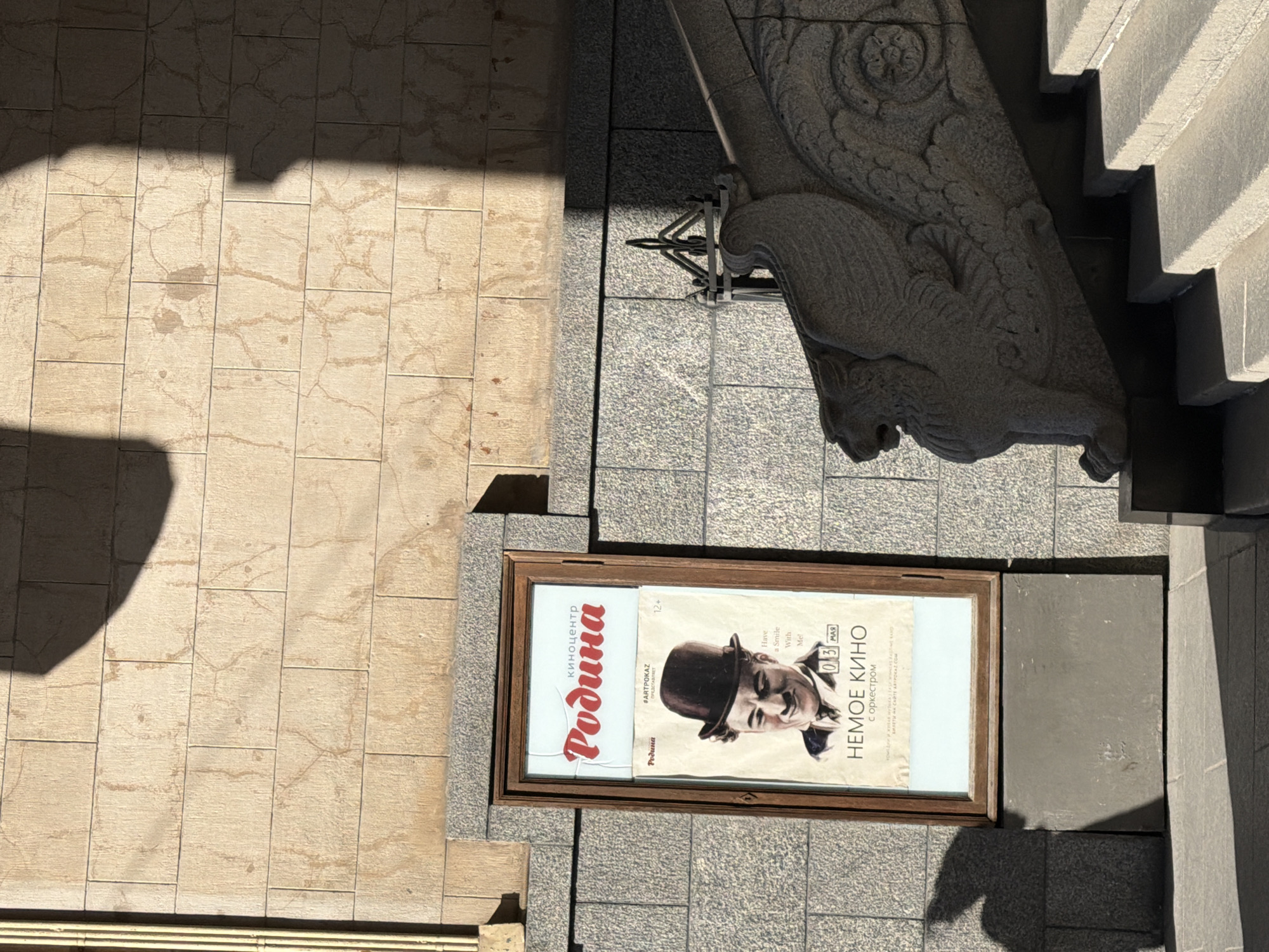 The image displays an outdoor, ground-level scene in Saint Petersburg, Russia, viewed from above, with strong, distinct shadows suggesting bright daylight. The primary subject is a wooden-framed advertisement board placed on dark grey, coarse-grained tiled pavement.
The advertisement poster inside the frame prominently features a black-and-white portrait of a man in a bowler hat, smiling, characteristic of Charlie Chaplin. Large red Cyrillic text at the top reads "киноцентр Родина" (Kinocenter Rodina), indicating the cinema venue. Below this, "#ARTPOKAZ ПРЕДСТАВЛЯЕТ" (ARTPOKAZ PRESENTS) suggests an event series. Other visible text includes an age rating "12+", an English phrase "Have a Smile With Me!", and the date "03 МАР" (March 3rd). The main event advertised is "НЕМОЕ КИНО с оркестром" (Silent Cinema with Orchestra).
To the left of the advertisement board, lighter, yellowish-brown tiled pavement extends, marked by varying textures and some discoloration. Prominent shadows from an unseen structure fall across this tiled area. On the right side of the frame, an ornate stone architectural element is visible, featuring intricate carvings, including what appears to be a winged mythological creature (possibly a griffin or gargoyle-like figure). Adjacent to this, a section of light grey textured steps or a stepped wall rises upwards. The scene captures a moment of bright, clear weather in an urban setting, likely near a cultural venue.