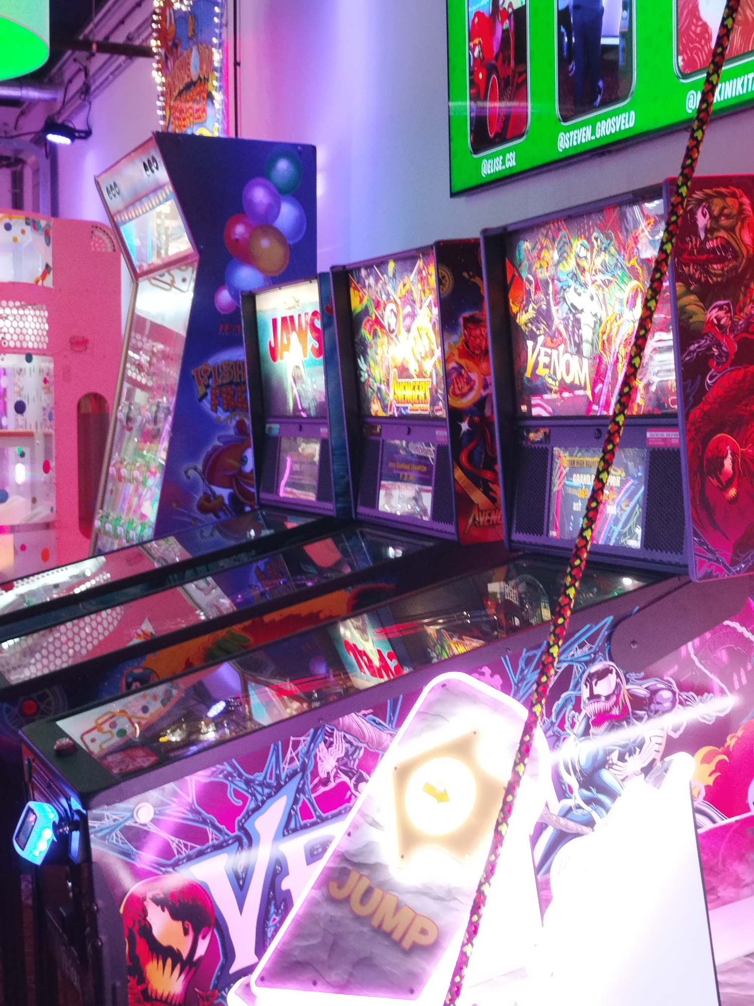 This media file shows a row of arcade games, likely pinball machines, in a dimly lit indoor setting. The machines are brightly lit with neon and LED lights, primarily in shades of pink, purple, and blue, creating a vibrant and somewhat futuristic atmosphere. The artwork on the machines features popular comic book characters, including Venom and other Marvel characters like Jaws and potentially Captain America.
One machine clearly displays the title "VENOM," with artwork depicting the character. Another features "JAYS" in bold lettering. The overall scene suggests a lively arcade or entertainment venue, possibly a bar or a dedicated arcade hall, given the ambiance. The reflections on the glass surfaces of the machines indicate a polished environment. The visible text on a screen above the machines includes social media handles like "@ELISE.CSL," "@STEVEN.GROSVELD," and "@KINIKIT." The presence of these social media tags might suggest the machines are part of an event or promotion. The cord of a game controller or accessory, with a red, yellow, and black braided pattern, runs diagonally across the image, adding a tactile element to the visual. The lighting is dramatic, with some areas appearing quite bright and others cast in shadow, highlighting the colorful graphics of the arcade games.
