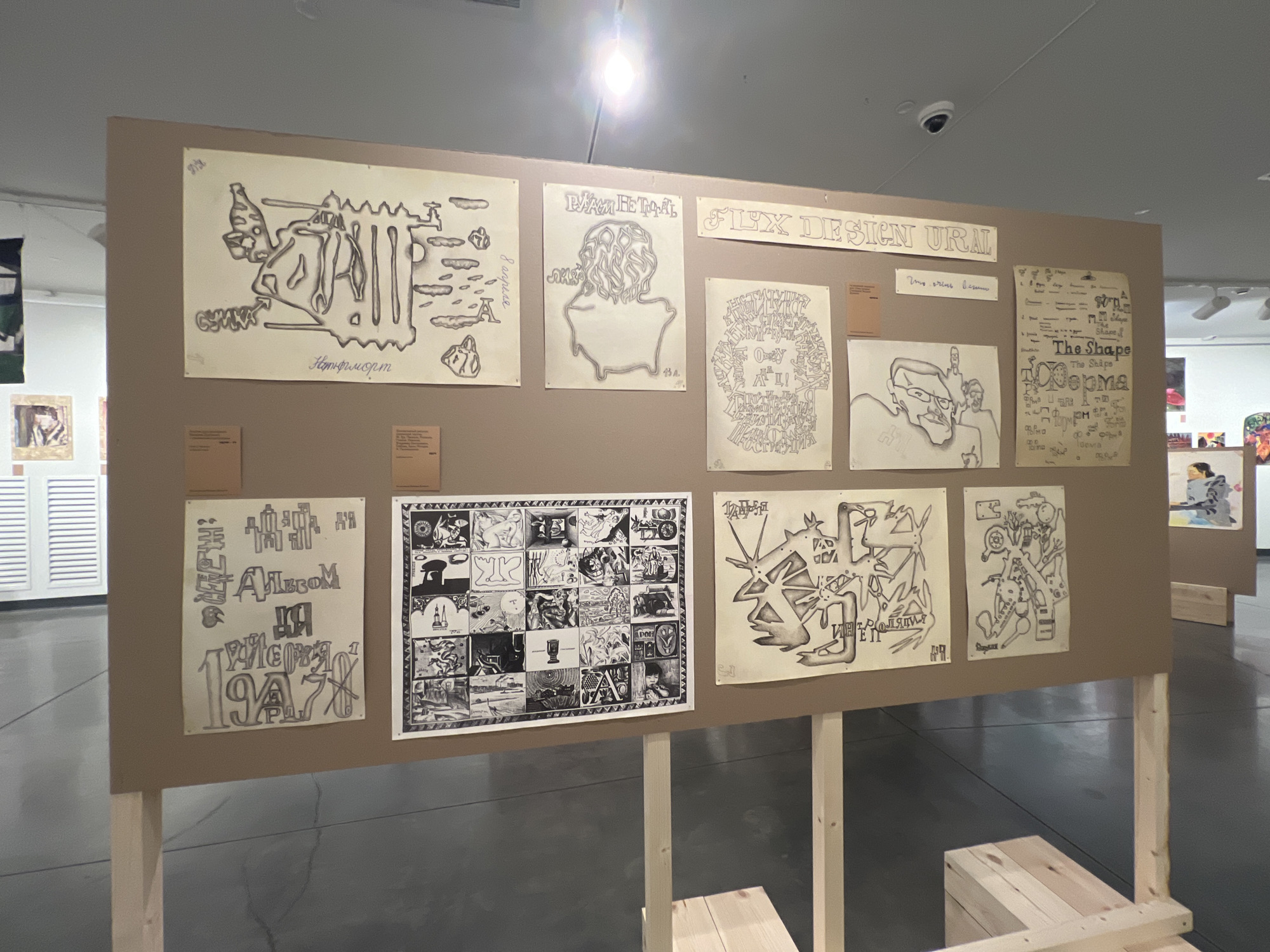 This image depicts an art exhibition in Yekaterinburg, Russia. The exhibition features a display of drawings and artwork mounted on a large, brown cardboard panel supported by wooden beams. The artwork includes a variety of styles, from abstract designs and caricatures to more complex, narrative-style illustrations.
The drawings are arranged in rows and columns. In the top row, from left to right, are several pieces. The first appears to be an abstract figure with text underneath, possibly in Russian. The second drawing shows a stylized head and shoulders with elaborate hair. The third is a banner-like piece with the text "FLUX DESIGN URAL" in a decorative font.
The second row contains more drawings. One is a complex, intricate composition with many small figures and shapes. Another is a portrait of a man with glasses, rendered in black and white. To the right of this portrait are two more abstract drawings.
The bottom row features a large grid of smaller illustrations, resembling a comic strip or a series of vignettes. To the left of this grid is a drawing with what appears to be stylized lettering and the date "1947". To the right, a single drawing with abstract figures and text.
The exhibition space appears to be a museum or gallery, with a polished concrete floor and neutral-colored walls. In the background, other artworks and displays are visible, as well as lighting fixtures and a security camera mounted on the ceiling. The overall impression is of a contemporary art exhibition showcasing diverse artistic expressions.