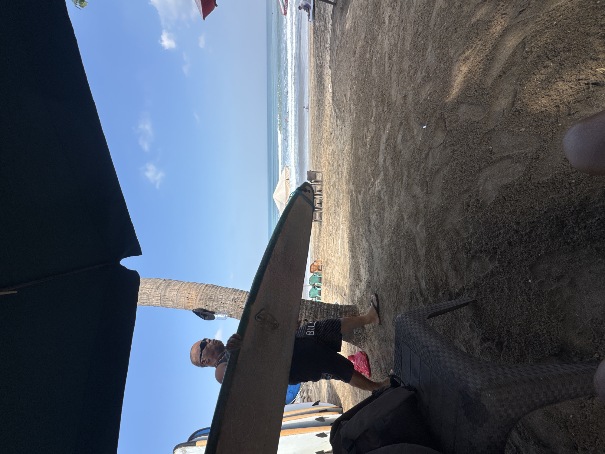 A man carrying a surfboard walks on the sandy beach in Kuta, Indonesia, under a blue sky. He is wearing sunglasses, a dark tank top, and black shorts. Behind him, other surfboards stand upright, and a palm tree is visible. In the distance, the ocean has waves, and several people are in the water surfing. Beach chairs and umbrellas are set up on the sand. The foreground shows a dark wicker chair with a backpack on it, and more sand.