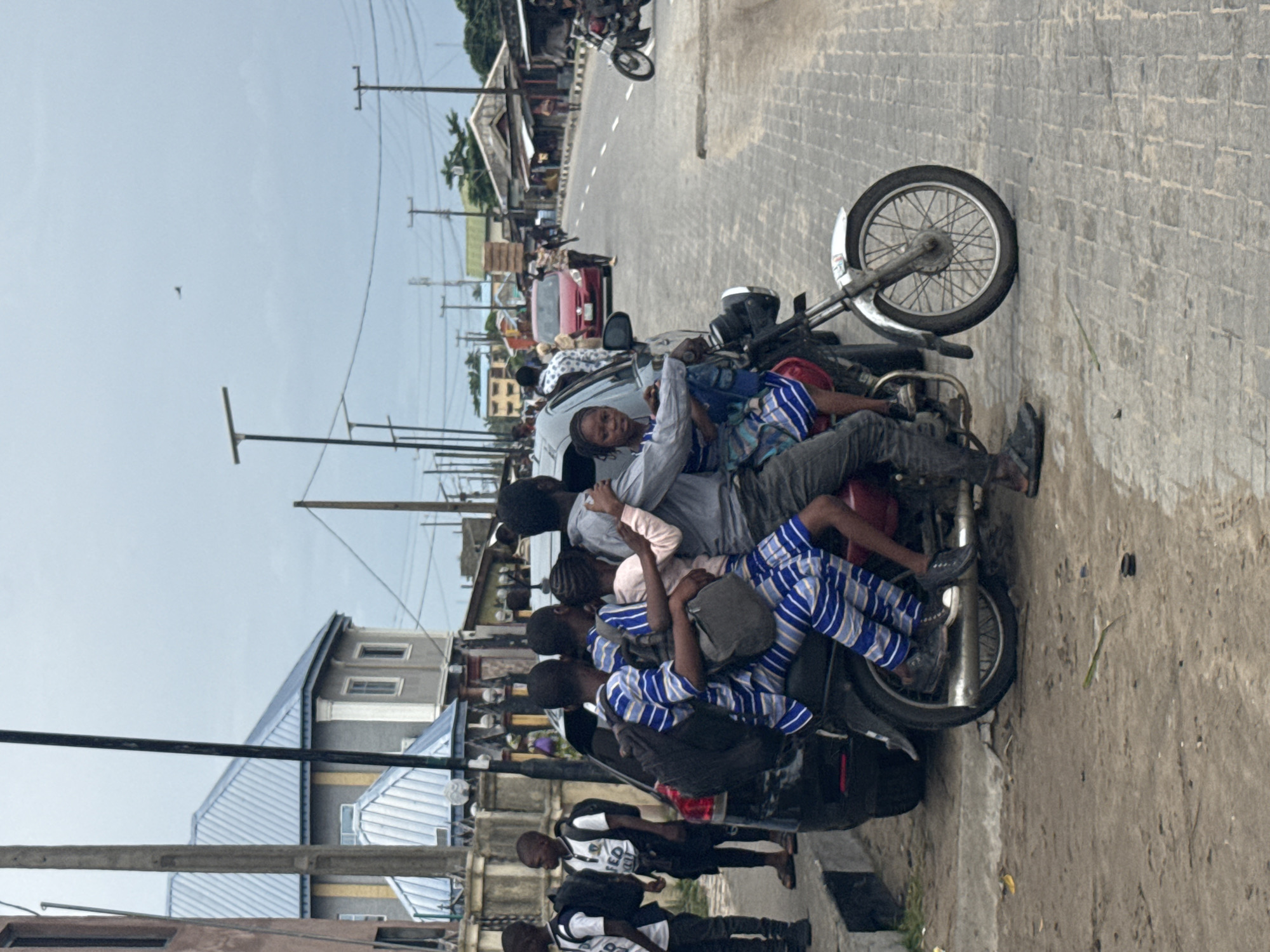 The image shows a group of people crowded onto a motorcycle in Badagry, Nigeria. Seven individuals are seen atop the motorcycle, with others standing nearby. The setting is a roadside in what appears to be a densely populated area, featuring buildings and utility poles, also other motorcycles and cars are present. The sky is overcast.