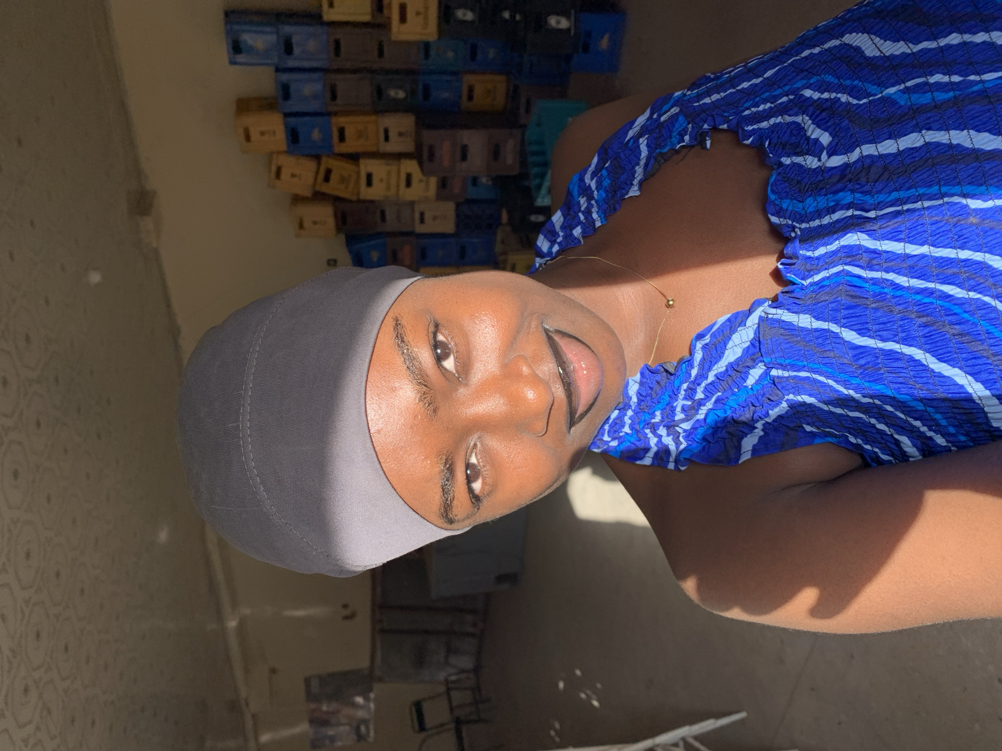 A young Black woman is depicted in a medium close-up shot. She is wearing a gray head covering and a blue and white striped top. A thin gold necklace is visible on her neck. The background shows a stack of multi-colored crates along a wall. The location is identified as New Stadium, Nigeria. A floor and possibly chairs are seen in the lower part of the image.