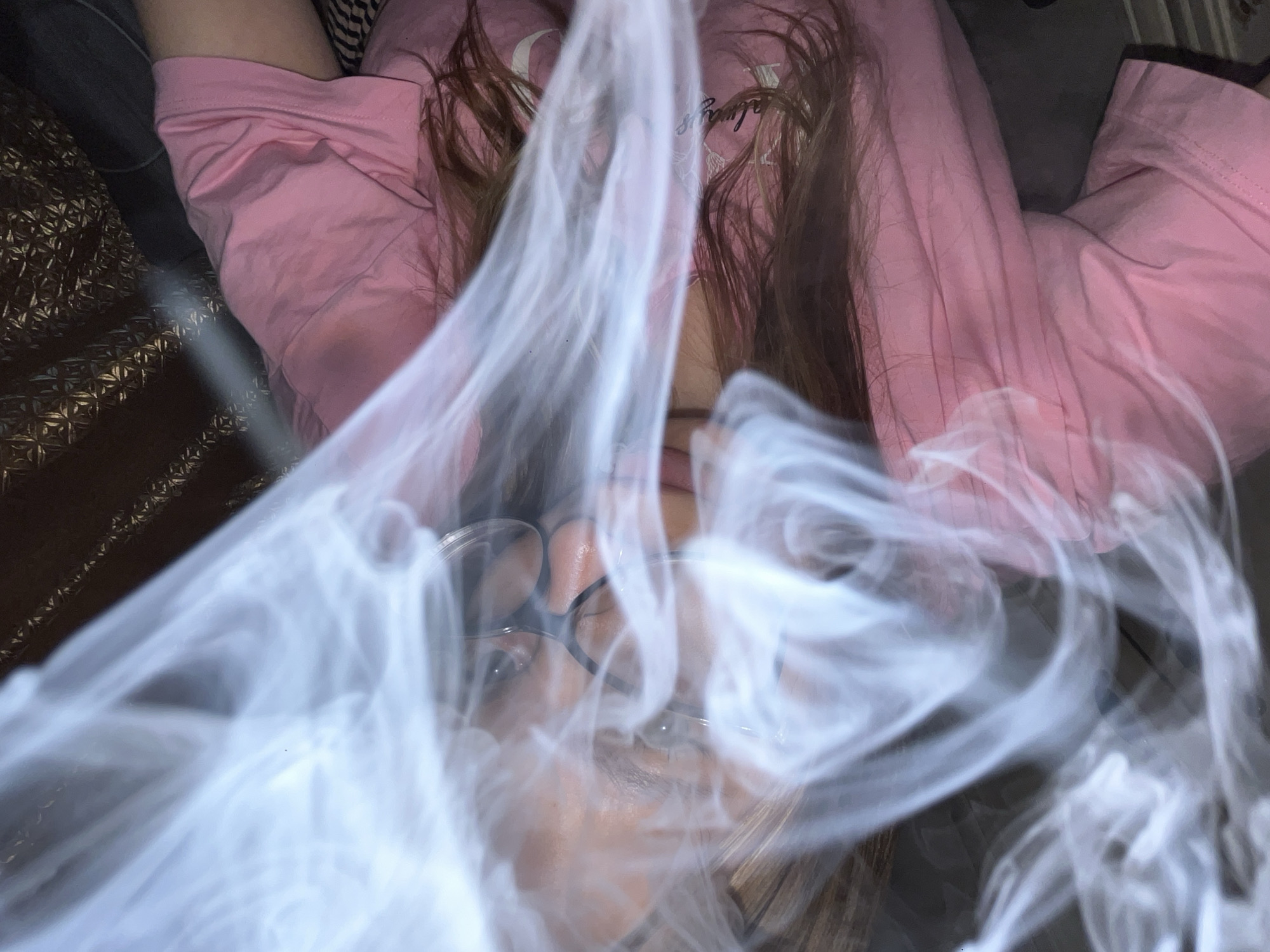 A person wearing glasses and a pink shirt is lying down with smoke obscuring their face. The person's brown hair is visible. The background includes a textured brown and gold surface, and a dark gray surface. The location is not discernible as Novopokrovskaya, Russia.