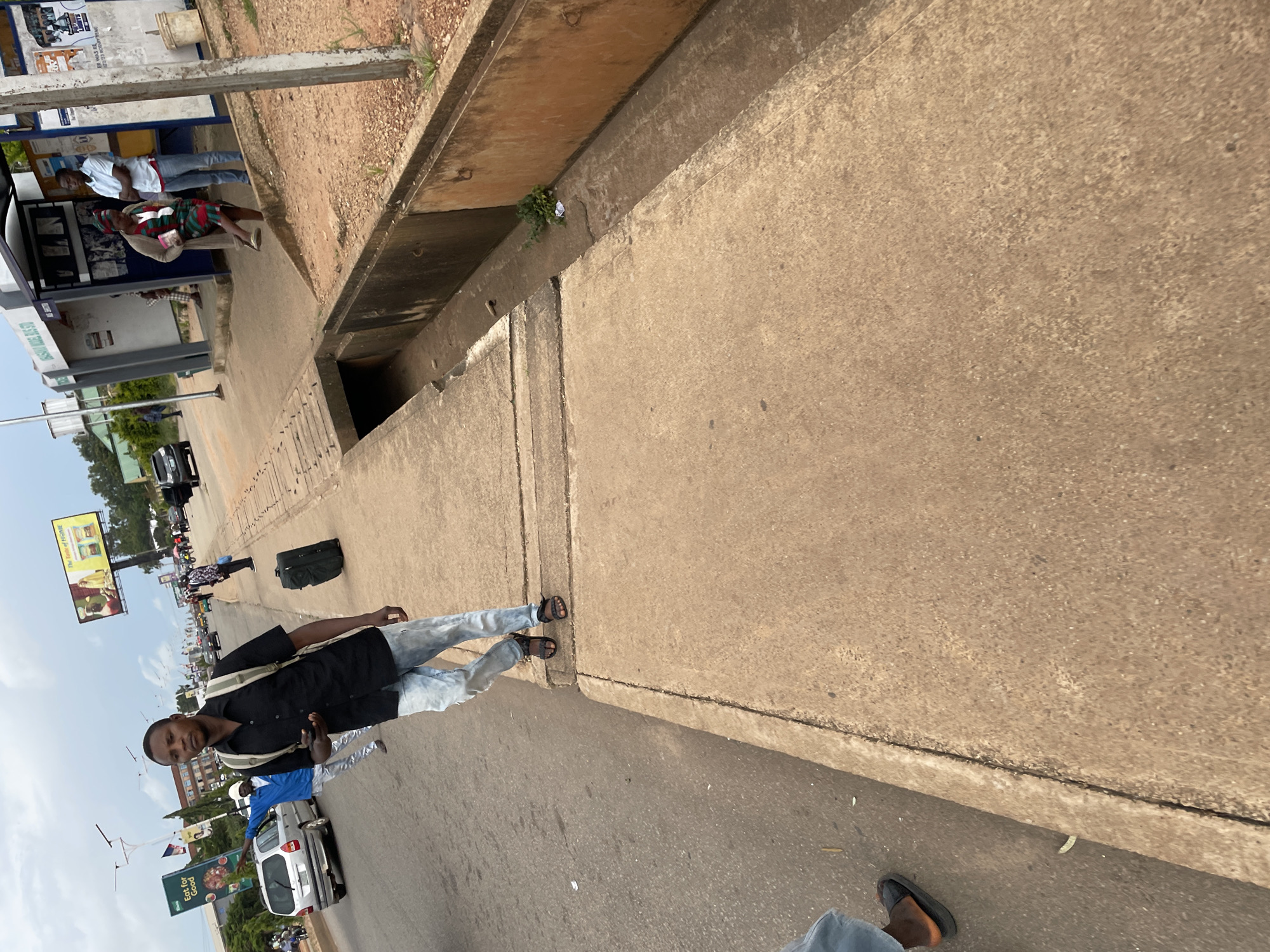 In Jos, Nigeria, an African man, identified as wearing a black shirt, light blue jeans, and sandals, walks along a paved sidewalk next to a busy road. He is carrying a black backpack and a black rolling suitcase. Other individuals are visible in the background, including a person in a blue shirt and another in a checkered skirt, standing at a bus stop. The scene includes a concrete drainage ditch running alongside the sidewalk, a bus shelter, streetlights, billboards, and several vehicles on the road, including a silver hatchback. The sky is overcast.
