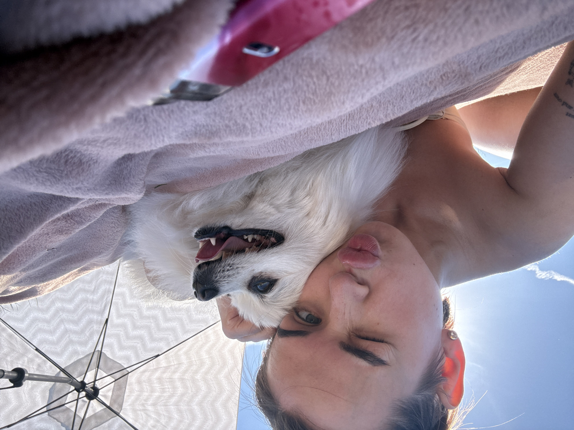 The image shows a woman and a white dog in an upside-down selfie. The woman is making a kissing face and winking one eye. The dog's mouth is open, appearing to pant. A light pink towel is draped over them, and an open white umbrella is visible below. The bright blue sky is visible in the background. The location is not discernible from the image, therefore Russia, Лобаново, can't be verified.