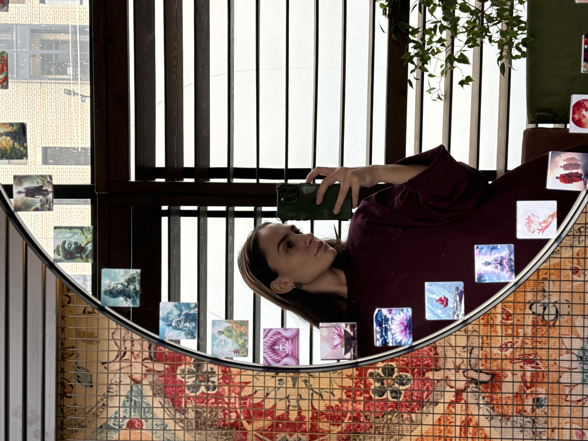 A woman is holding a smartphone, presumably taking a photo in front of a large, round mirror, which is adorned with small, square pictures. The mirror is set against a backdrop of vertical wooden slats that appear to be part of a balcony. In the background, a building is visible through the slats. The mirror itself is set upon a base with a pattern reminiscent of floral tiles or an intricate tapestry. This image could be taken in the restaurant "Franky Woo" in Krasnoyarsk, Russia.