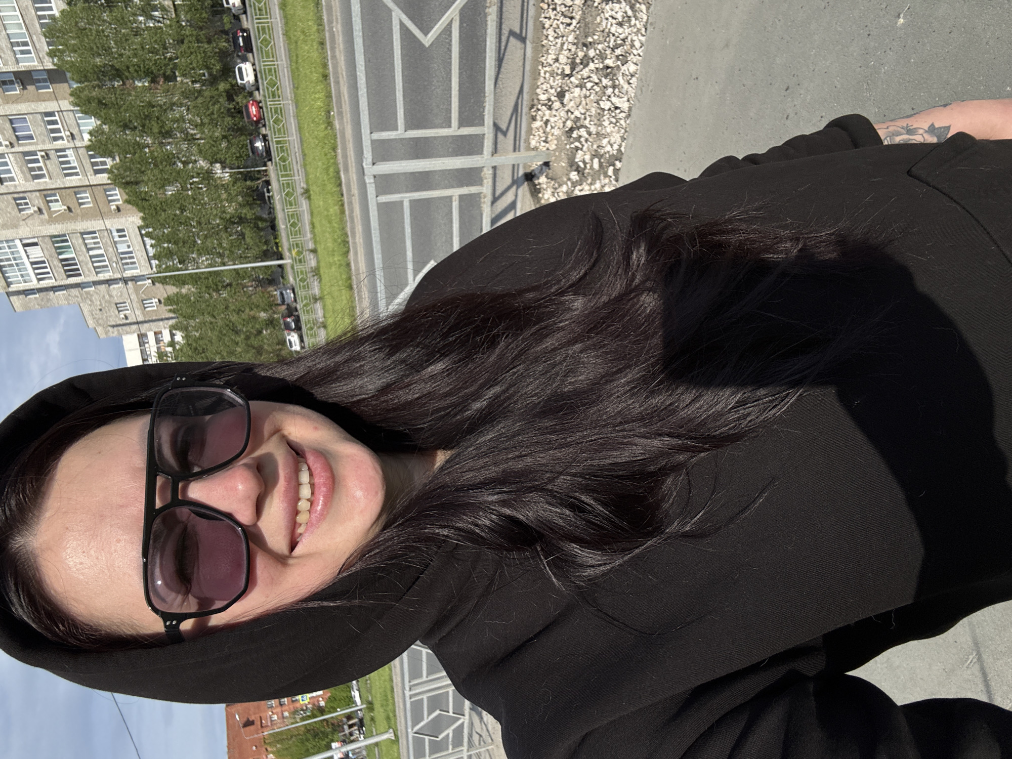 The image shows a woman wearing a black hoodie and sunglasses, smiling at the camera. The background includes a cityscape with buildings, trees, and a road with cars. The image appears to be taken outdoors, possibly on a sidewalk or pedestrian area.