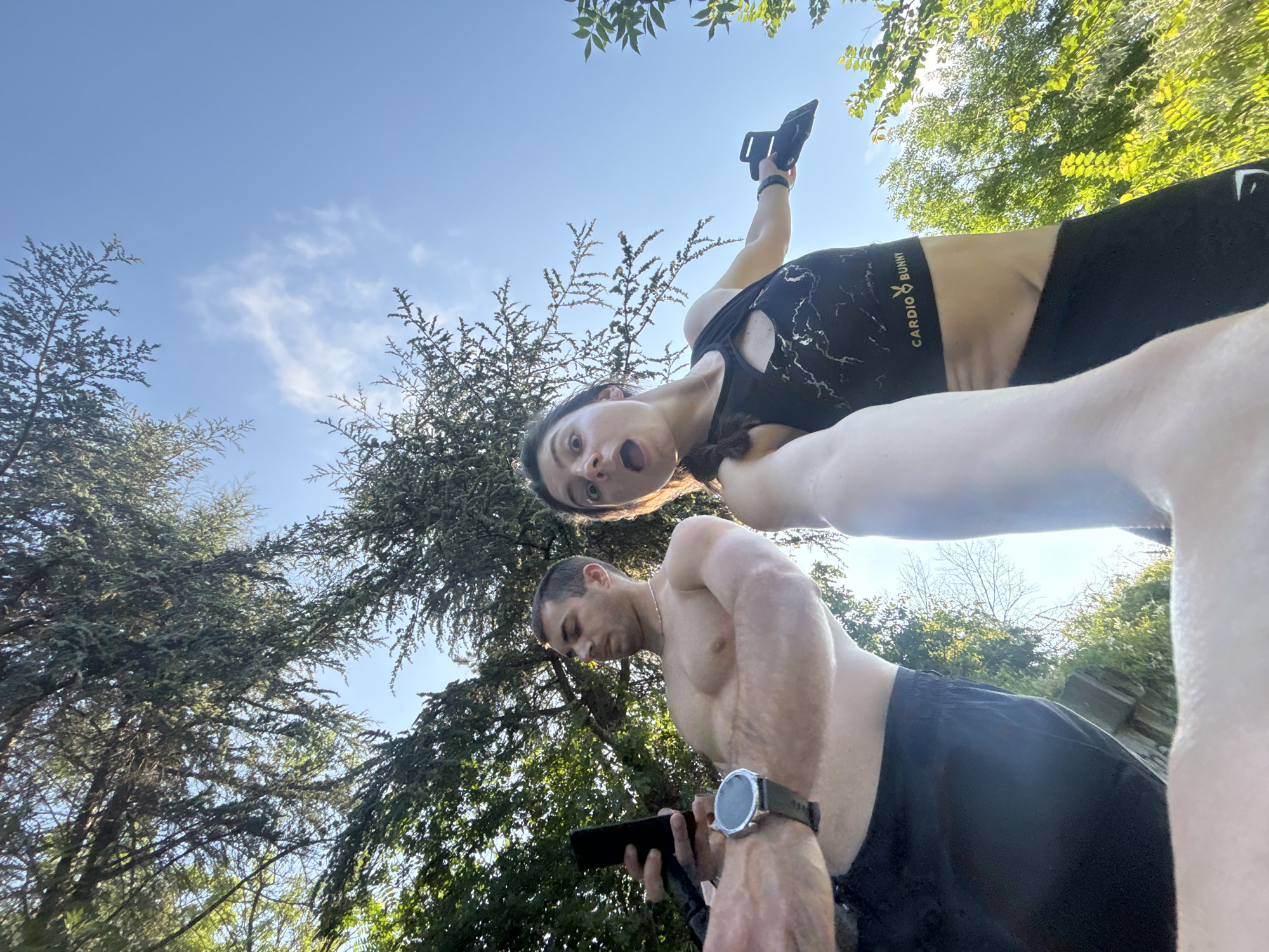 In a low-angle outdoor selfie, two individuals are visible against a bright blue sky with scattered clouds and lush green trees. The male subject, on the left, is shirtless and wearing black shorts. He holds a smartphone in his left hand and wears a smartwatch on his left wrist. His gaze is directed at the phone. To his right, the female subject, also in athletic wear, has a surprised expression. She wears a black sports bra with a cutout design and black leggings. She holds a black object, possibly a phone holder or accessory, in her right hand. The background consists of leafy branches of trees and foliage, suggesting a park or outdoor recreational area. The overall impression is that of a fitness-related activity or a casual outdoor moment. The location is not explicitly identifiable within the image itself to be Varna, Bulgaria.