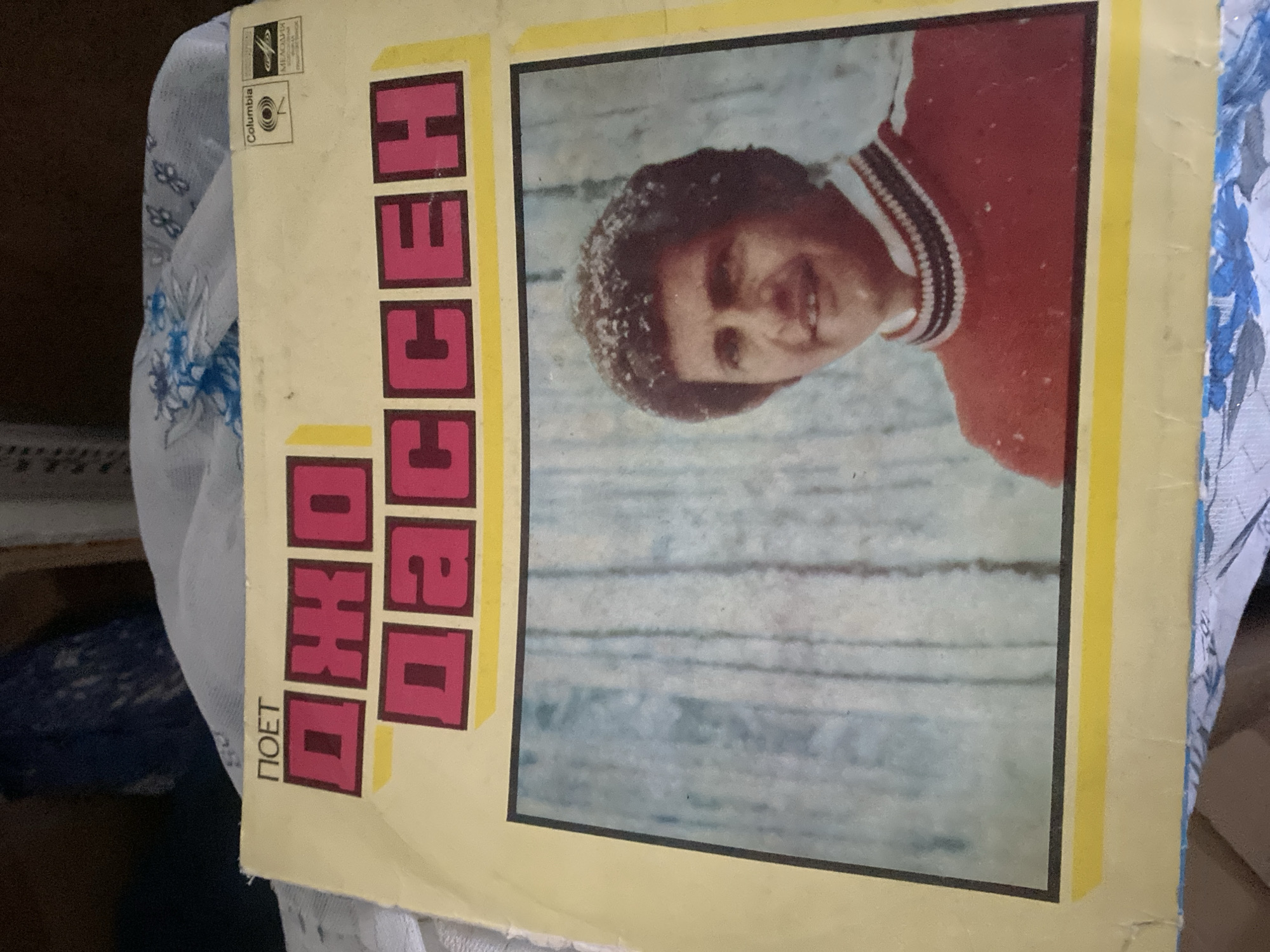 This is a close-up shot of a vintage record album cover. The album is yellow with a black border framing a photograph of a man. The man has dark, curly hair and is smiling. He is wearing a red sweater with a white and black striped collar. The background of the photograph appears to be a winter scene with a light blue and white textured surface, possibly snow-covered trees or fabric. The title of the album is printed in large, bold, pink block letters with a yellow outline. Above the title, in a smaller black font, it reads "ПОЕТ". In the upper right corner, there are two logos: one for Columbia and another, a circular logo with text that is partially obscured. The album is lying on a surface covered with a patterned fabric, possibly a blanket or tablecloth, which has blue and white floral motifs. The overall impression is a well-preserved but aged record album. This object is not in Nizhny Novgorod, Russia.
