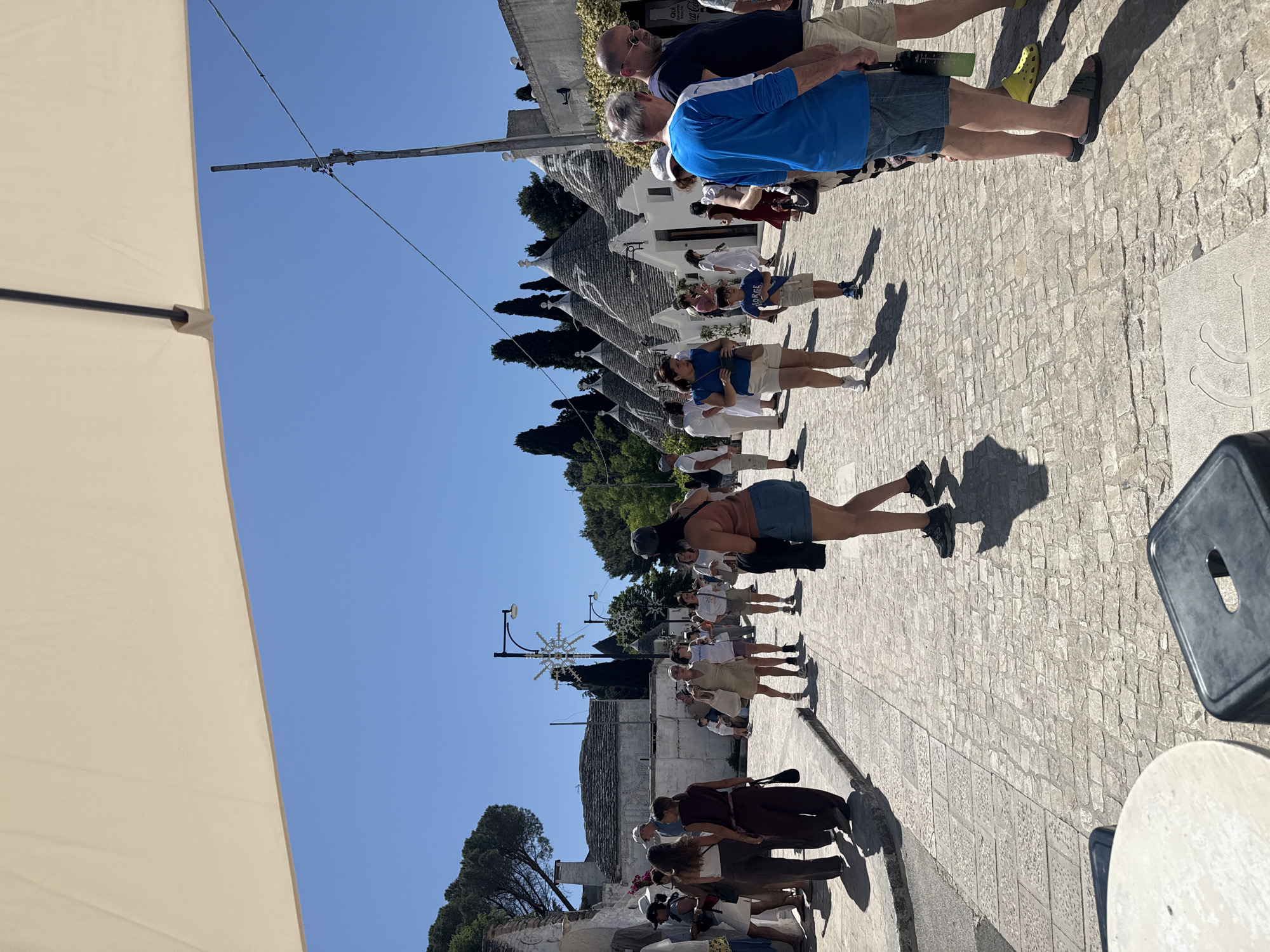 The image shows a street scene in Alberobello, Italy, populated with a line of tourists. The iconic Trulli houses, with their conical roofs, are visible along the street. The tourists are standing on a paved surface, with an umbrella shading part of the scene. Some of the tourists are touching each other. Some are wearing casual clothes, including shorts and t-shirts. Clear blue sky.