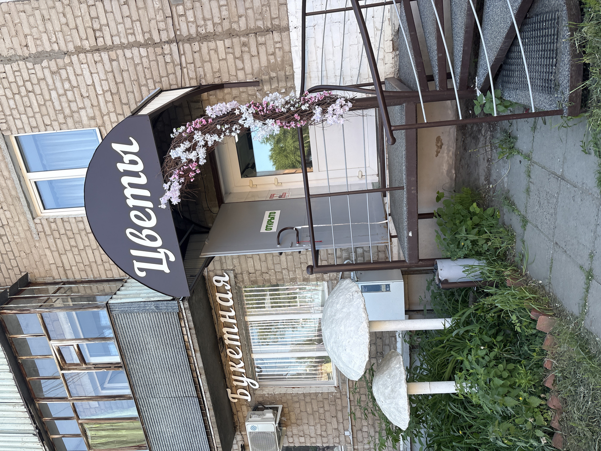 The image shows the exterior of a flower shop, located in Chaikovsky, Russia. The sign above the shop reads "Цветы" (Flowers) and "Букетная" (Bouquet). The entrance is decorated with a floral wreath. There are two mushroom-shaped decorations in the foreground. The building is brick, with a window and an air conditioning unit visible. Stairs lead up to the entrance. Greenery is present on the ground.
