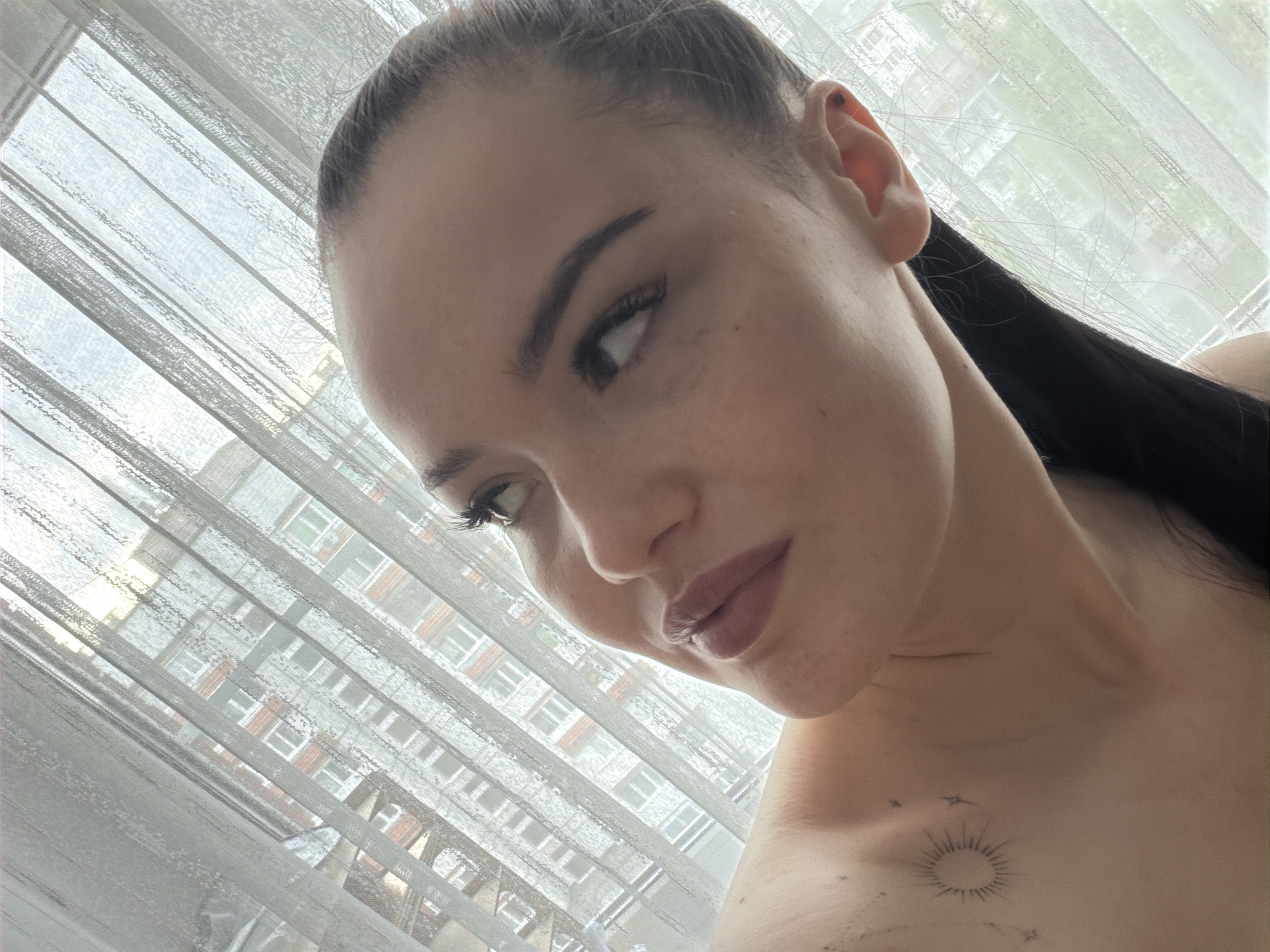 A close-up portrait of a light-skinned woman with dark hair pulled back, positioned in front of a window with white blinds. Her gaze is directed downwards and to the left. She has dark eyebrows and eyelashes, and freckles are visible on her face. Her lips are a muted pink shade. On her upper chest, a tattoo depicting a sun or starburst with accompanying smaller star-like elements is visible. Through the window blinds, a multi-story building with numerous windows is visible. The building's architecture suggests that the location is likely in the city of Chaikovsky, Russia. The overall lighting is soft, diffused, and dominated by a white-ish tone.