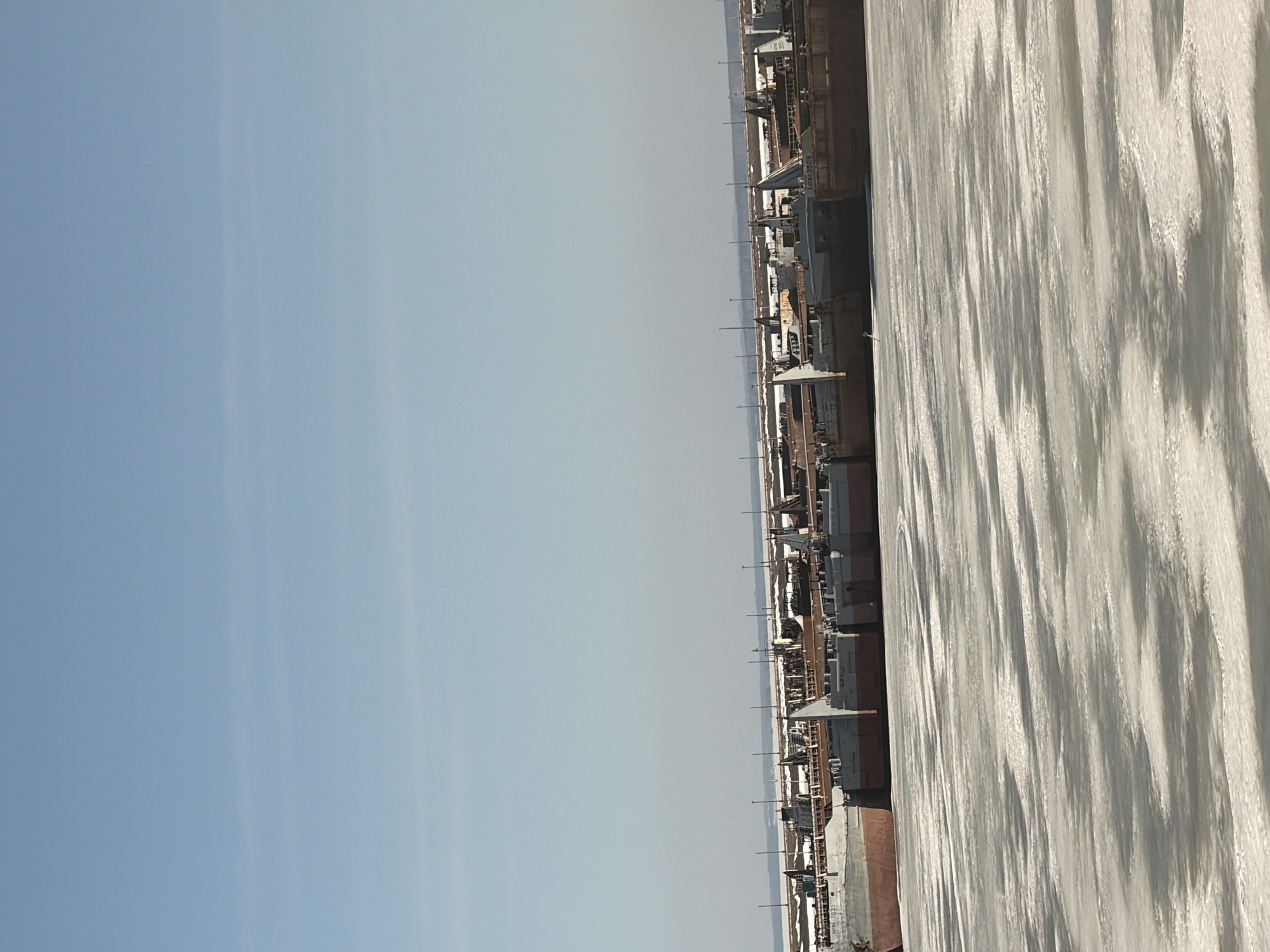 The image shows the Kama River in Chaikovsky, Russia, covered in ice. Along the riverbank are moored ships or barges. The sky is clear and blue.