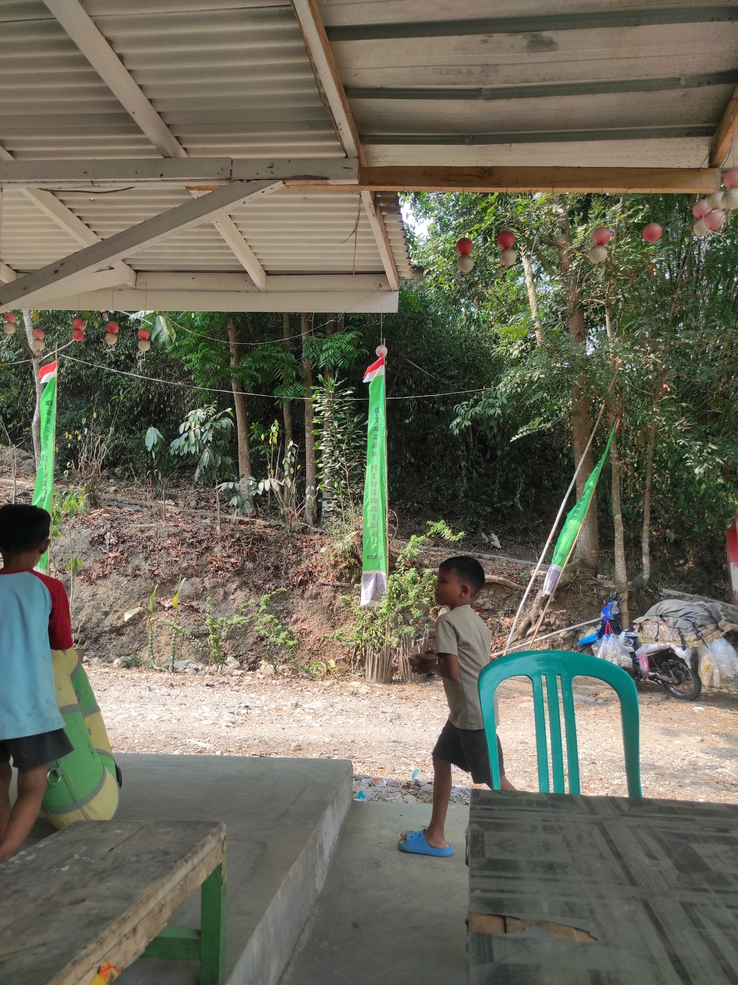 A medium shot captures two young boys standing in an outdoor area with a covered roof. The boy on the left, facing away from the camera, wears a blue shirt with red sleeves and black shorts, and a large green and yellow object is beside him. The boy on the right, facing left in profile, wears a tan t-shirt and black shorts, and blue flip-flops. He is walking towards the left side of the frame. Behind them, a lush green, heavily wooded hillside slopes upwards. Two long green banners with white text and red and white flags are prominently displayed on poles in front of the hillside. A turquoise plastic chair is visible in the foreground, and a wooden table is partially in view at the bottom of the frame. A motorcycle is parked in the background on the right side, covered with plastic bags. The ground is a mix of dirt, gravel, and scattered debris. The overall setting appears to be a roadside stall or rest stop in Mekarwangi, Indonesia, under a corrugated metal roof supported by white beams.