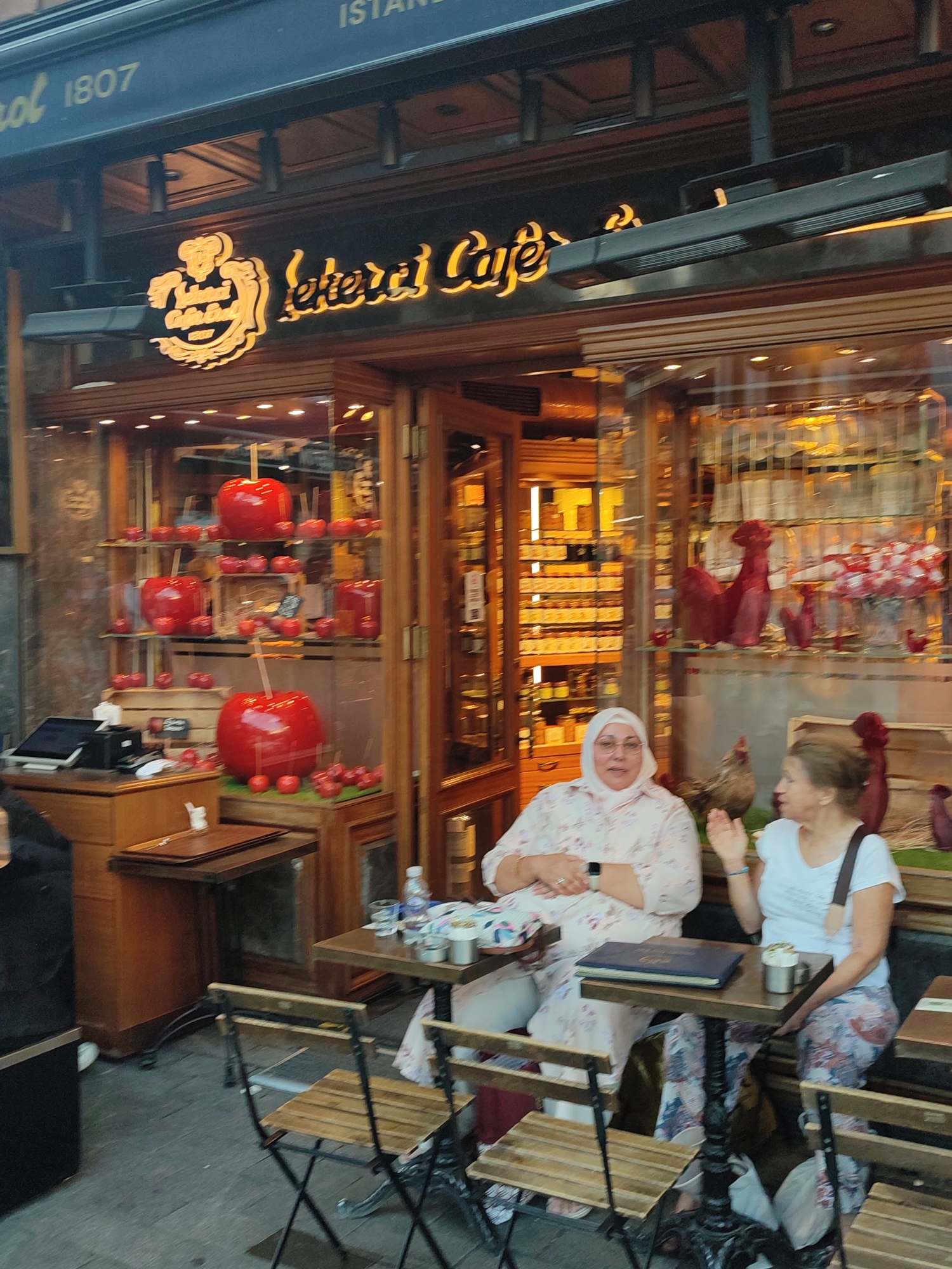 The image captures a storefront in Kadıköy, Turkey, with a sign reading "Lateur Cafe" and "1807". Two women are seated at a table outside the cafe. The woman on the left wears a floral patterned headscarf and shirt, while the woman on the right wears a white t-shirt. They appear to be engaged in conversation. The cafe's window display features a prominent display of large, red, candy apples, along with smaller ones. Inside the cafe, shelves are stocked with various jars and products. The outdoor seating area consists of small wooden tables and chairs. The overall setting suggests a bustling street scene in a commercial district.