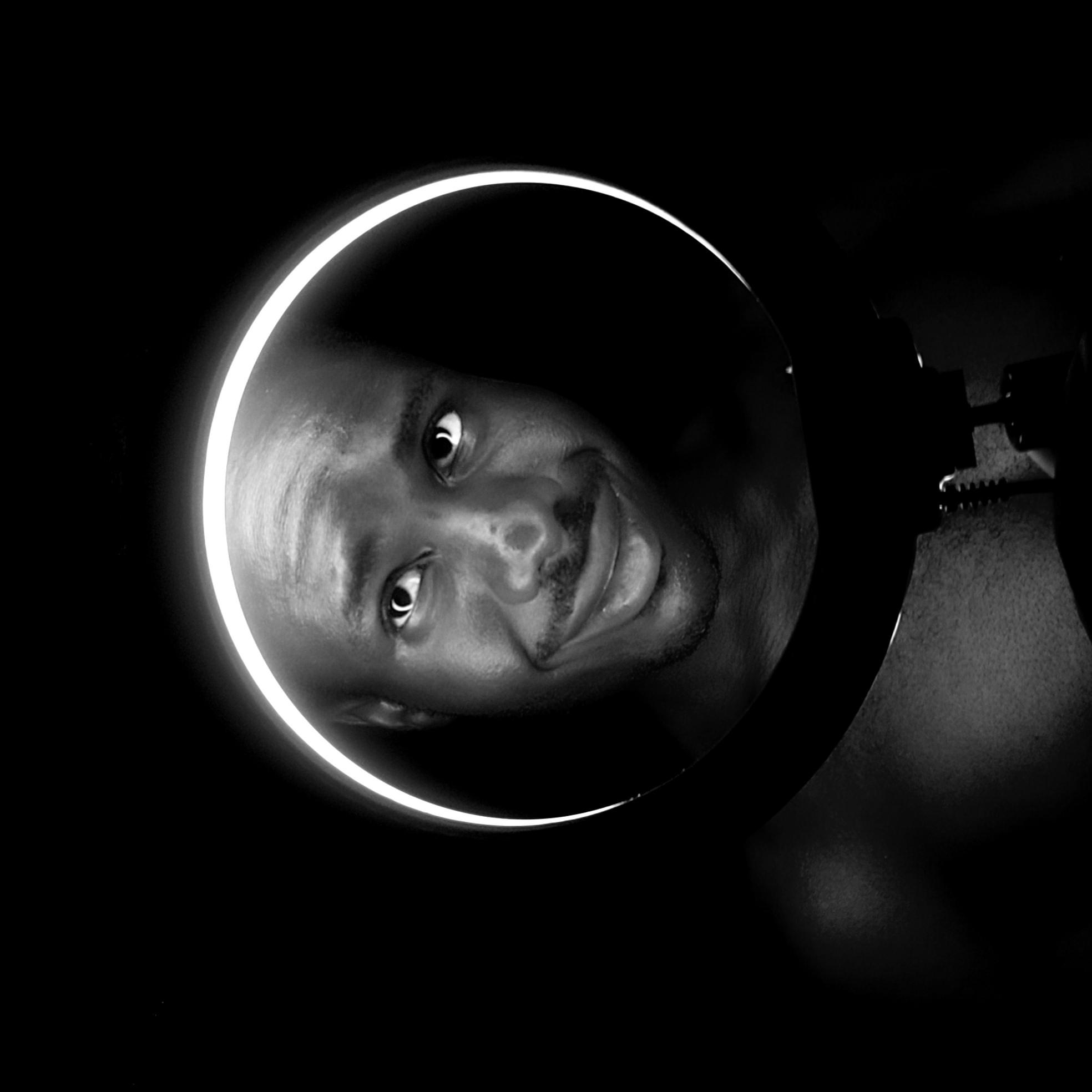 A black and white image shows a man’s face peering through a circular light. The man, who appears to have dark skin, is tilting his head to the left. His expression includes a slight smile. The circular light source is positioned around his face, illuminating it against the dark background. The setting appears to be indoors. Narayi, Nigeria is not determinable from the image.