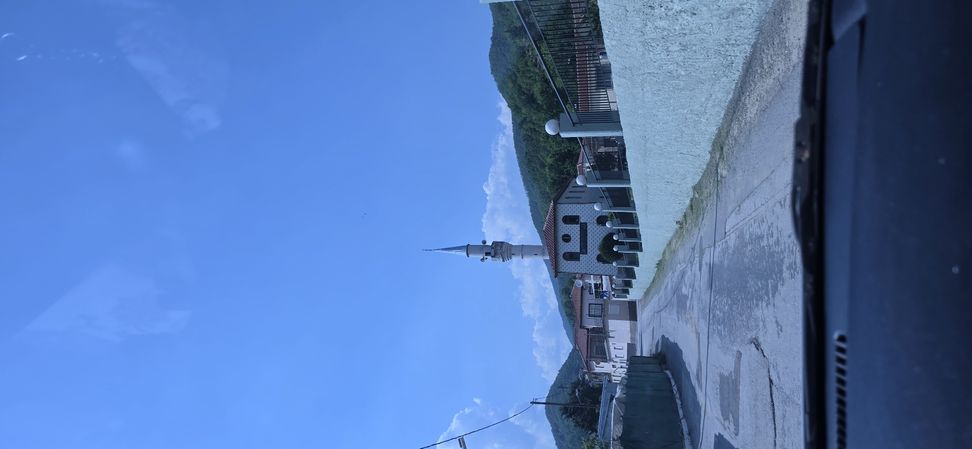 The image depicts a rural village scene, identified as Melivoia, Greece, captured from inside a vehicle. The foreground shows the top edge of a dark car dashboard and a windshield wiper base. A paved road, appearing to be asphalt or concrete with visible cracks and patches, extends into the midground, sloping gently upward and curving to the left. Along the right side of the road, a light greenish-blue stucco wall runs parallel, topped by a dark metal fence with regularly spaced white spherical finials. Behind this wall is a mosque, a prominent white building with a terracotta-tiled roof and several arched windows, featuring a tall, slender minaret with a light blue conical spire. To the left of the road, a green mesh or tarp partially obscures an indistinct structure. In the background, lush, tree-covered mountains rise under a bright blue sky dotted with scattered white cumulus clouds. Faint reflections, consistent with light sources or objects inside a vehicle, are visible in the upper sky portion of the image. Two small dark specks are discernible in the sky above the minaret.