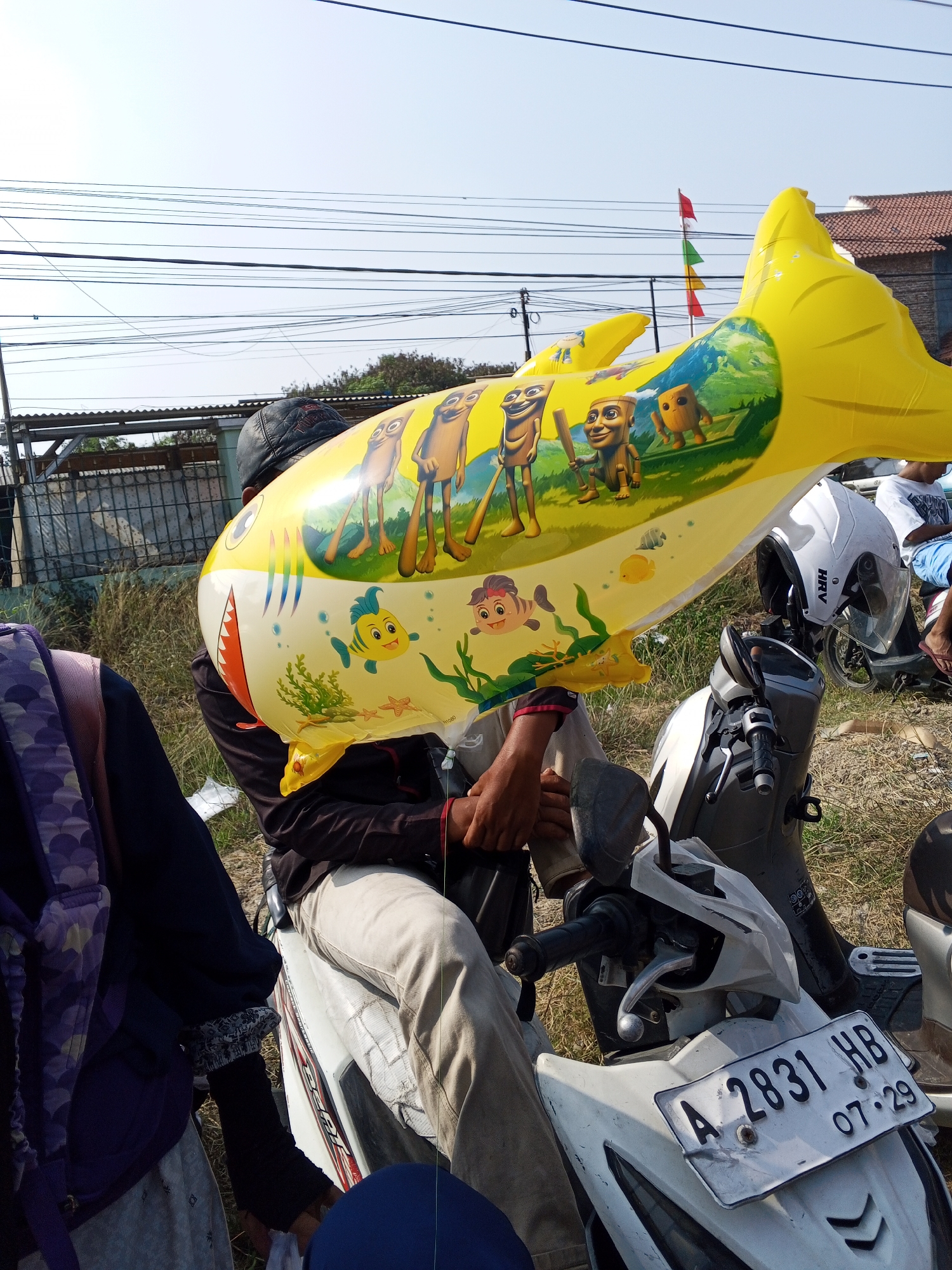 In Ranjeng, Indonesia, a person wearing a black cap sits on a white scooter, holding a large, deflated yellow inflatable shark. The shark is decorated with cartoon characters and images of marine life. The person is wearing a black jacket and light-colored pants. Another person, wearing a purple backpack, is partially visible to the left. A white helmet with the "HRV" logo sits on the scooter's handlebar. A white motorcycle with a license plate reading "A 2831 HB" is parked in front of the person on the scooter. The background shows a grassy area, trees, a building with a tiled roof, and power lines against a clear sky.