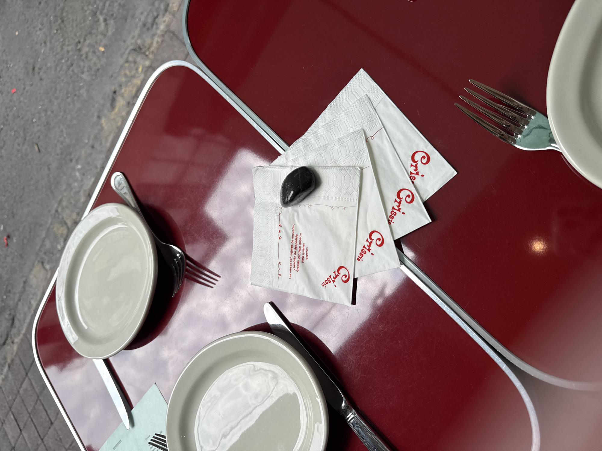 This image captures a scene at Crissis in Mexico City, Mexico. The focus is on a two-tiered, burgundy-colored table set for dining. On the table, there are three plates accompanied by silverware – knives and forks. Three Crissis-branded napkins are visible, with a smooth, dark pebble resting on top of one of them. The table is set outdoors, with a glimpse of the sidewalk pavement in the upper left corner. The lighting is bright and appears to be natural daylight.
