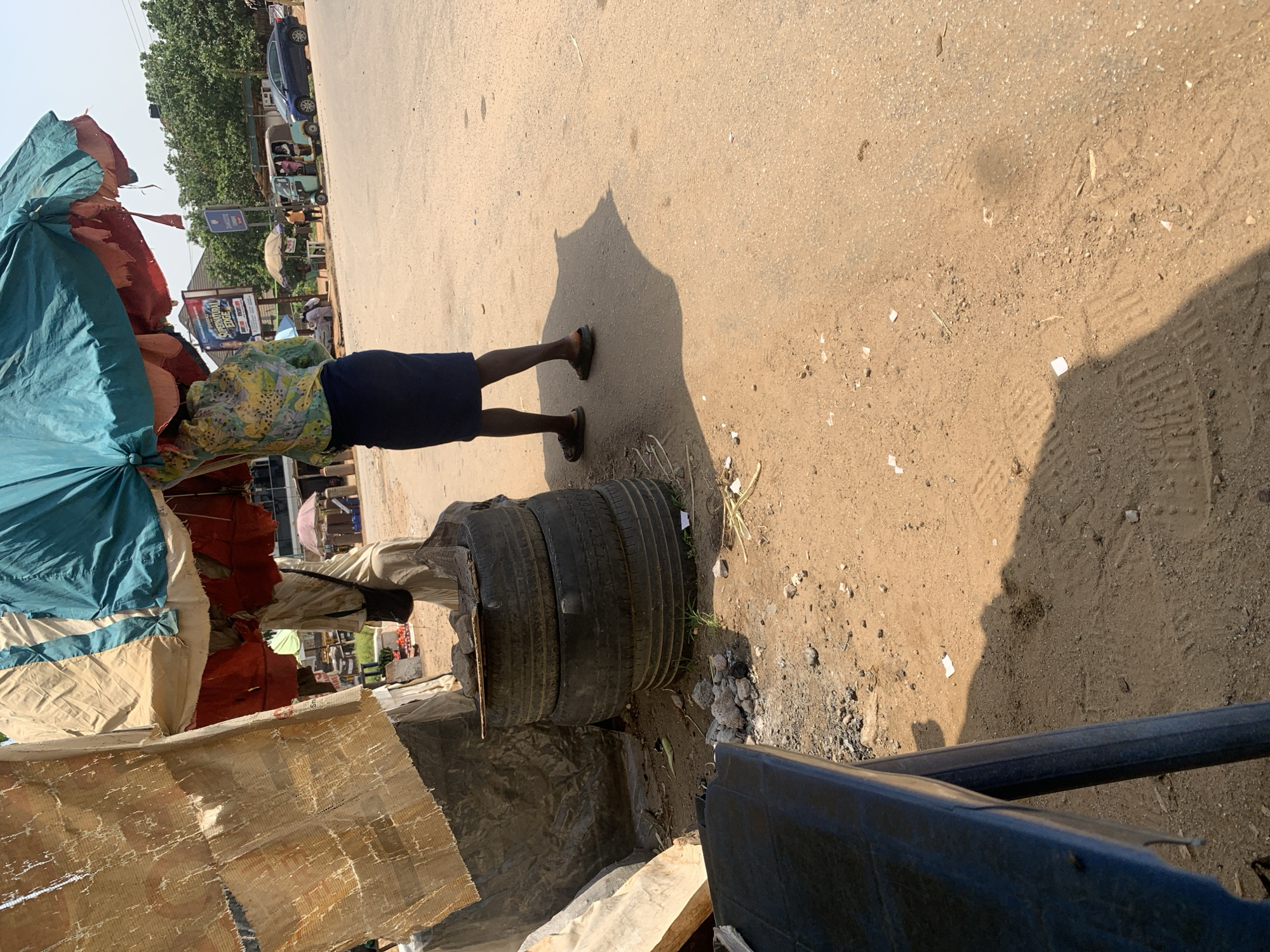 This image shows a street scene in Abuja, Nigeria. In the foreground, a person is standing under a large, multi-colored umbrella, holding it overhead. They are wearing a patterned long-sleeved shirt and a dark skirt. To the left of the person, there is a stack of three old tires. Behind the tires, a rough-looking structure with torn fabric is visible. The ground is covered in reddish-brown dirt and gravel. In the background, there are some market stalls with umbrellas, a few vehicles, and trees lining the street. The sky is clear and bright.