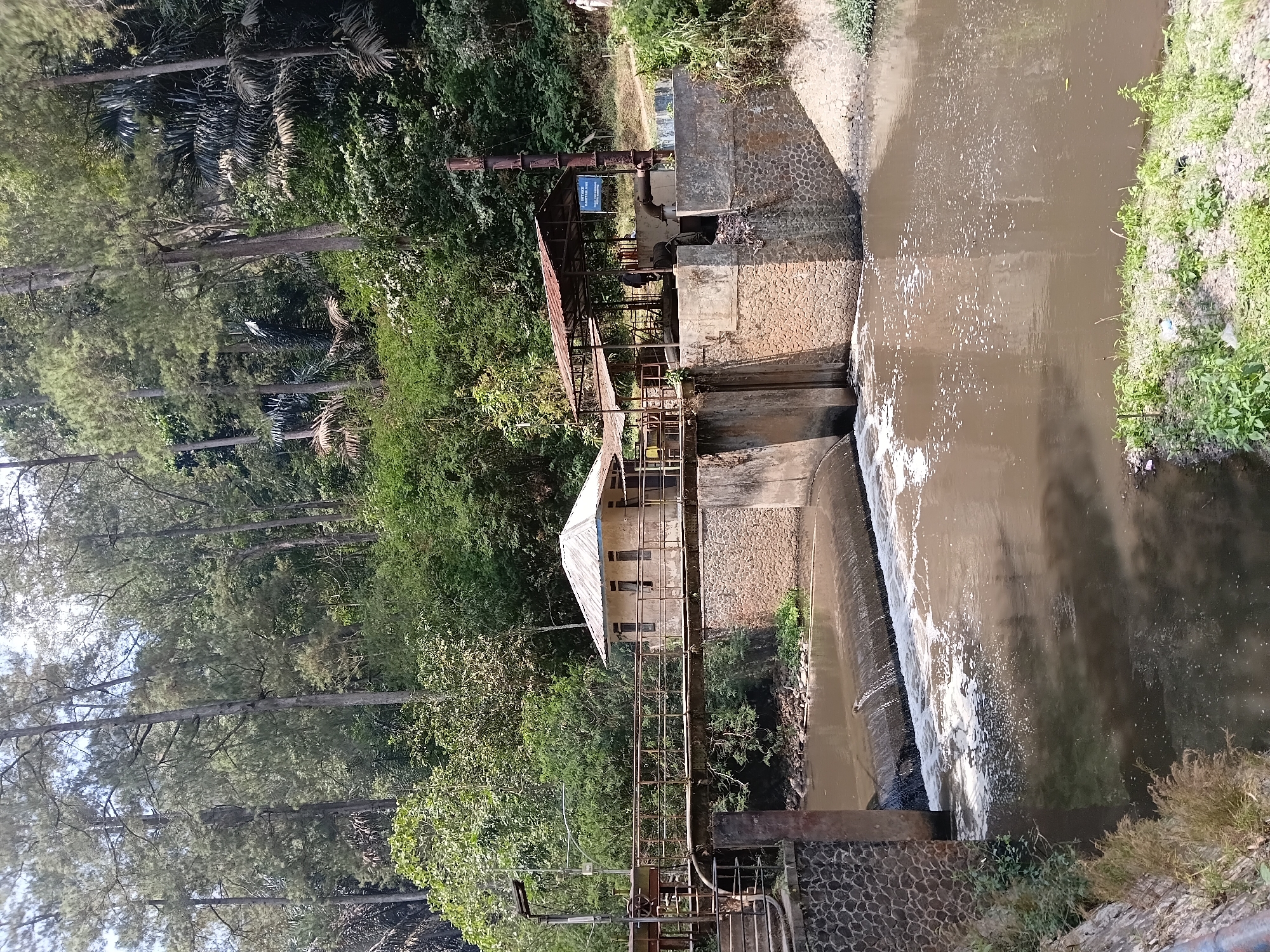 The image shows a small hydroelectric power plant situated in a forested area near the Ciburial river in Ciburial, Indonesia. A concrete dam with a stone facade has water flowing over it, creating whitewater. A small, light-colored building with a metal roof is located on top of the dam, connected by a rusty metal walkway. Adjacent to this building is an open-sided structure with a corrugated metal roof, housing industrial equipment. A tall, rusted metal pipe extends vertically from this area. The surrounding environment is lush with dense green foliage and tall trees. The river water appears murky. Two individuals are partially visible near the open-sided structure, and another person stands further in the background on the right side of the image. The scene suggests a functional, albeit somewhat aged, infrastructure within a natural setting.